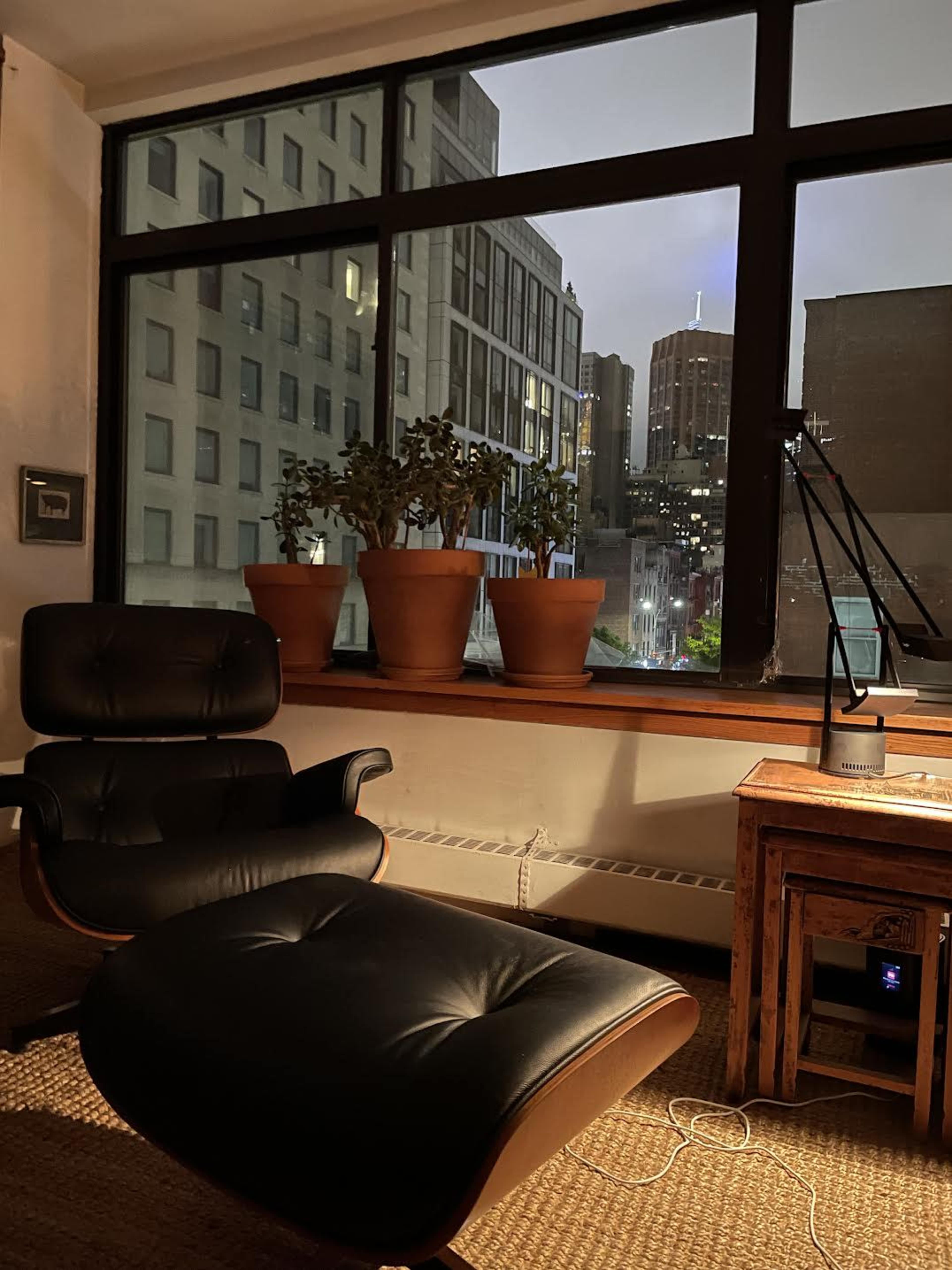 A dark leather chair and ottoman sit beside a wooden table, with potted plants on the windowsill overlooking a cityscape at night.