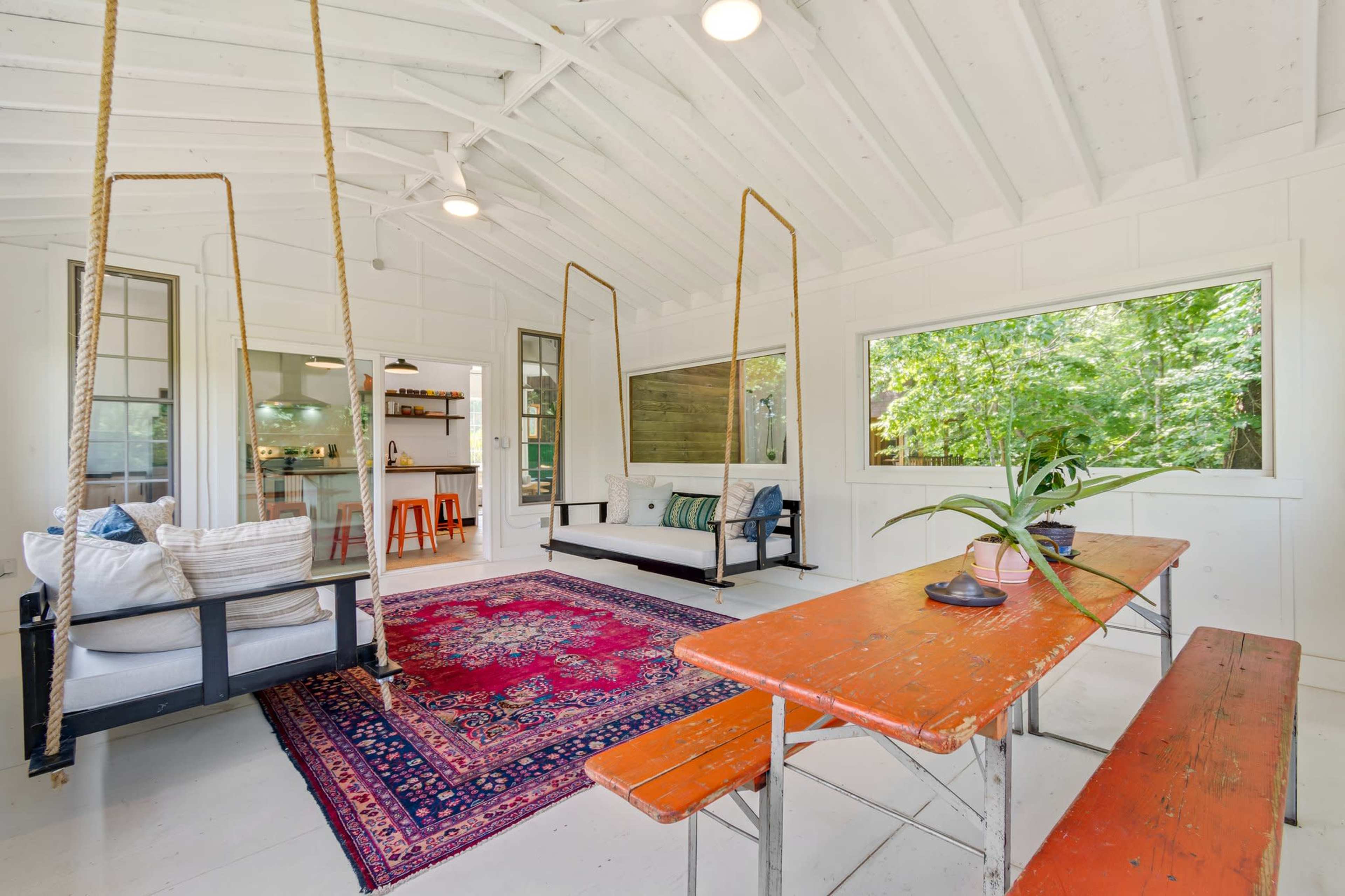 The interior of a bright, open space features a rustic table, hanging swings, and large windows overlooking greenery.