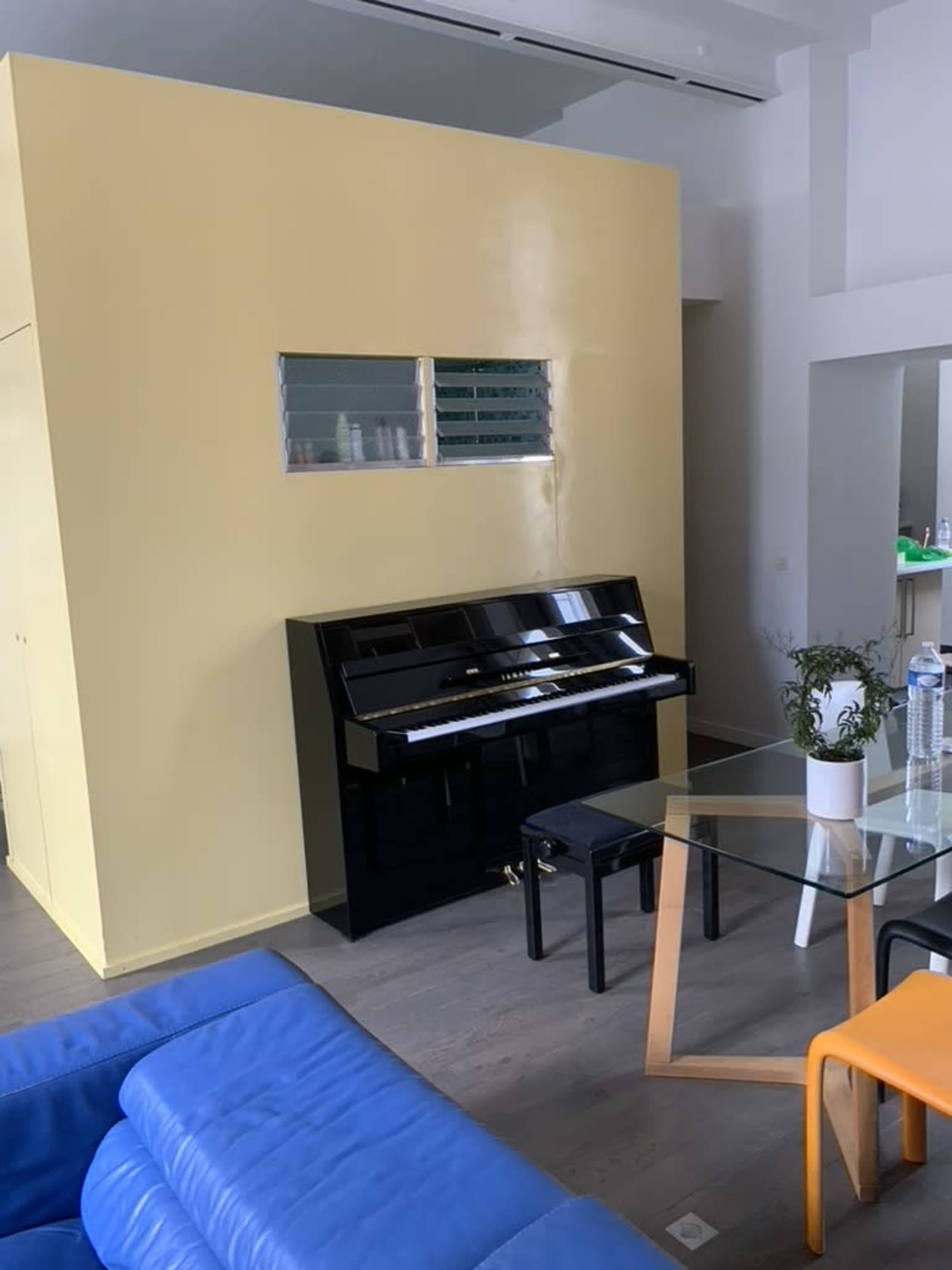 A black piano sits against a yellow wall in a room furnished with a glass table and blue and orange chairs.