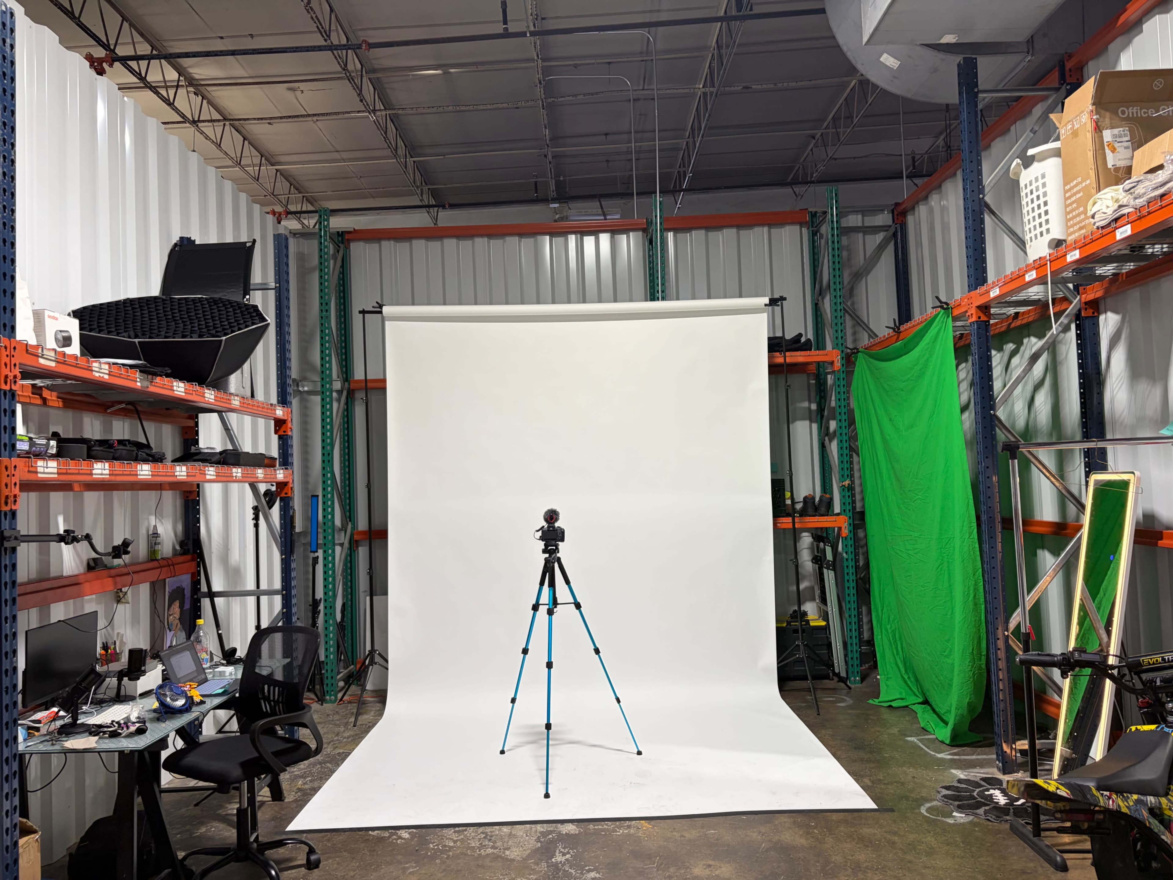 The image shows a photography studio setup featuring a white backdrop, a tripod with a camera, and cluttered shelving with various equipment on the sides.