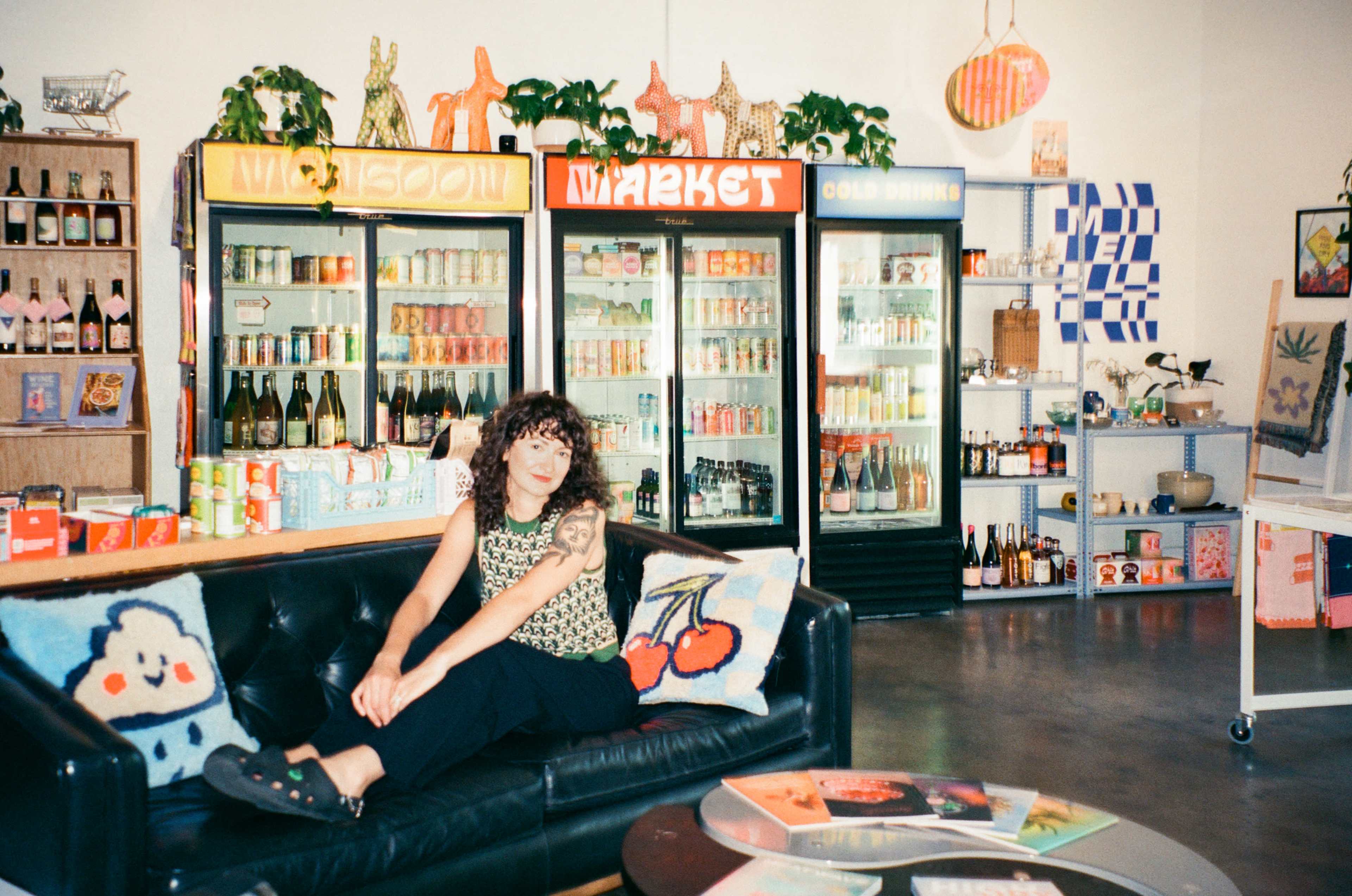 A person sits on a couch in a colorful market shop surrounded by shelves and refrigerators filled with various drinks and snacks.