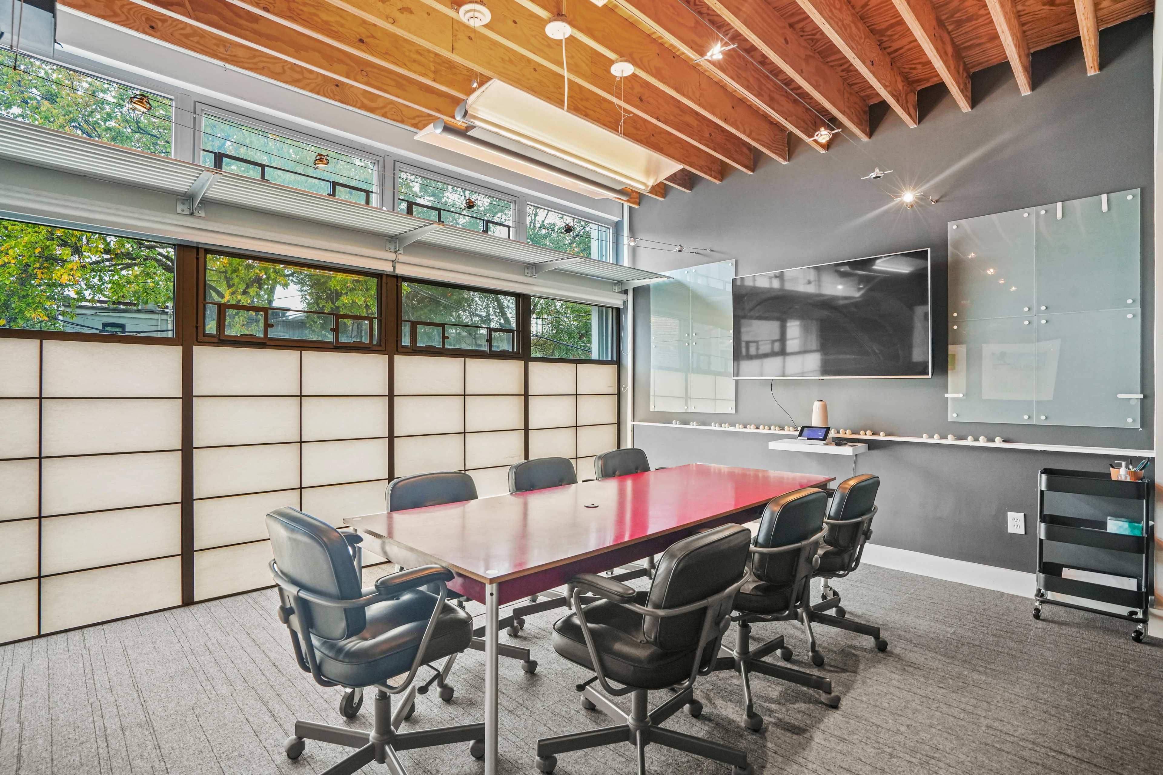 A modern conference room features a large table surrounded by black office chairs, with a wall-mounted screen and large windows allowing natural light to enter.