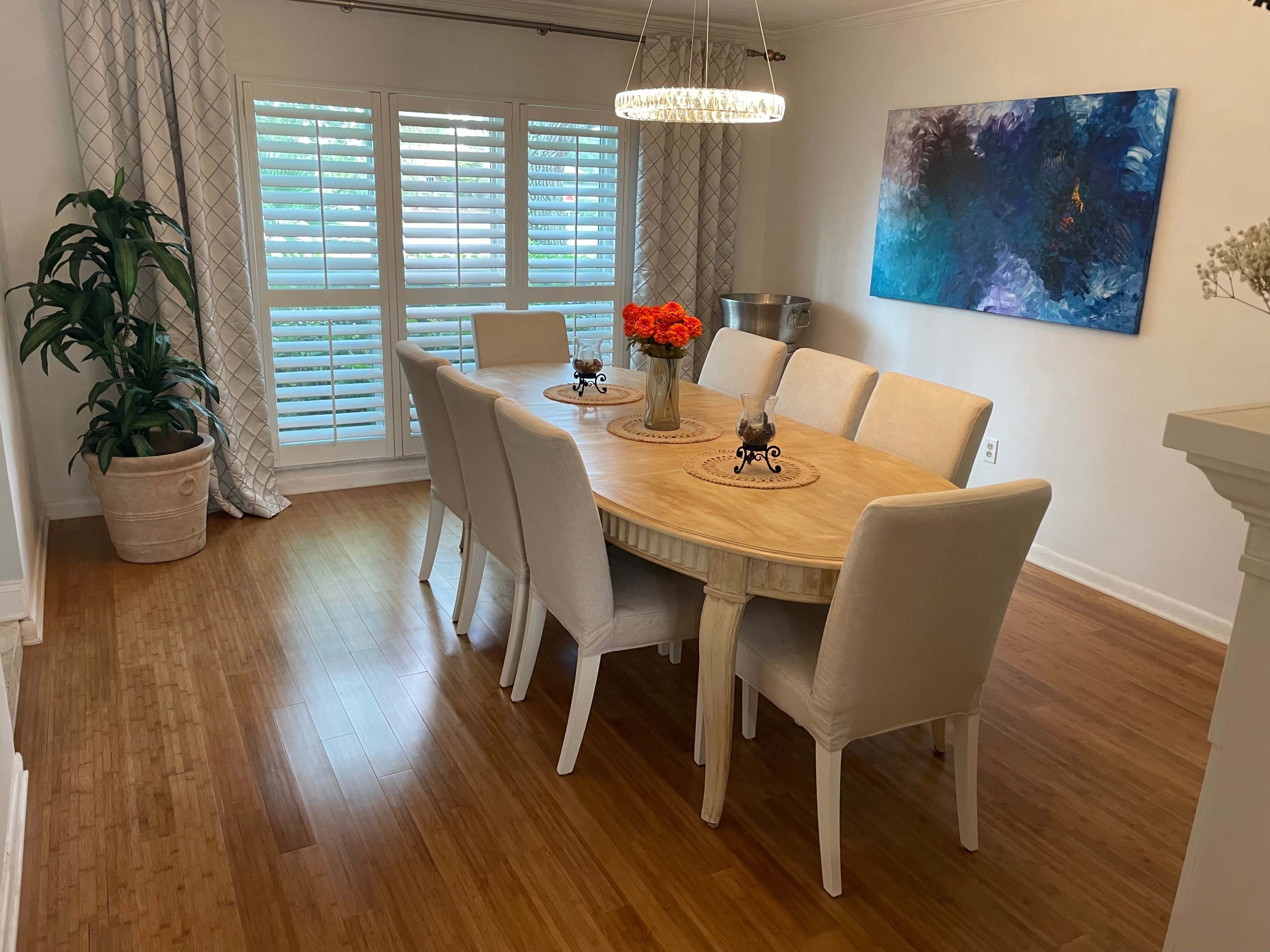 A dining room features a round wooden table with eight upholstered chairs, surrounded by large windows covered with white shutters and adorned with a colorful painting on the wall.