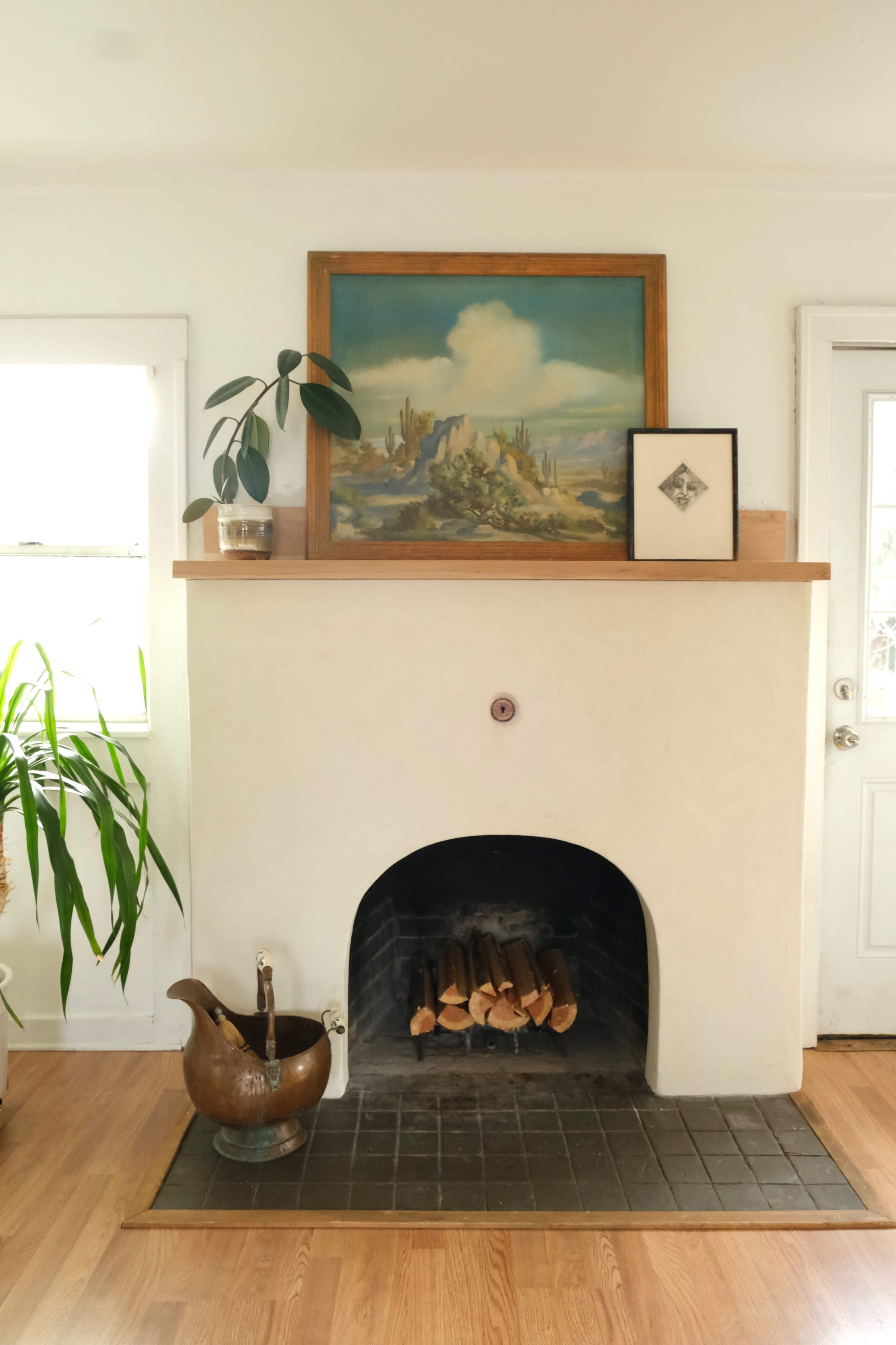A whitewashed fireplace with a wooden mantel displays a landscape painting, a framed artwork, and a potted plant, beside a door and a green plant in the corner.