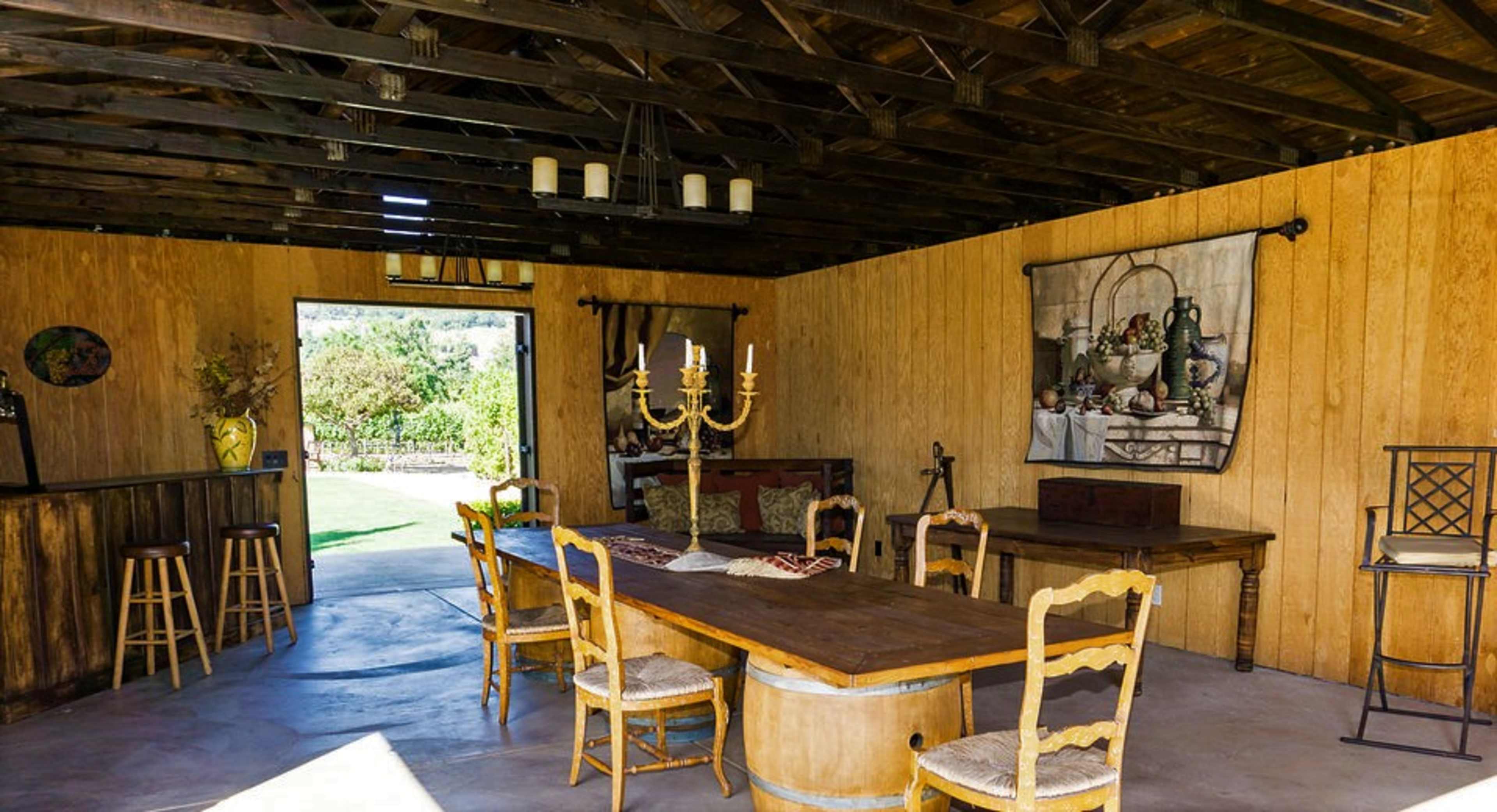 A rustic dining area with a large wooden table surrounded by chairs, a candelabra in the center, and an open doorway leading to a garden view.