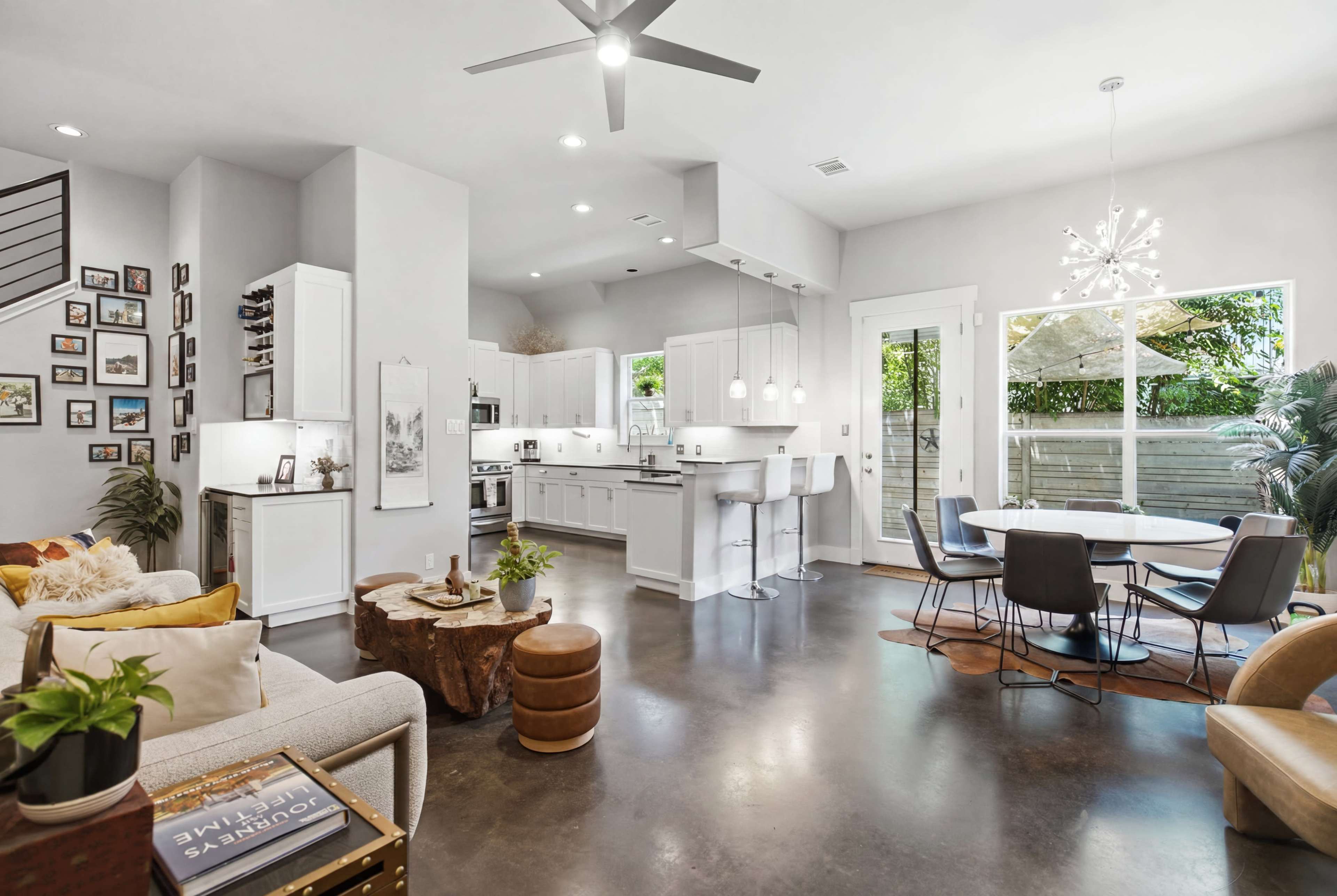 The image shows a modern open-concept living space featuring a kitchen, dining area, and seating area with large windows and polished concrete flooring.