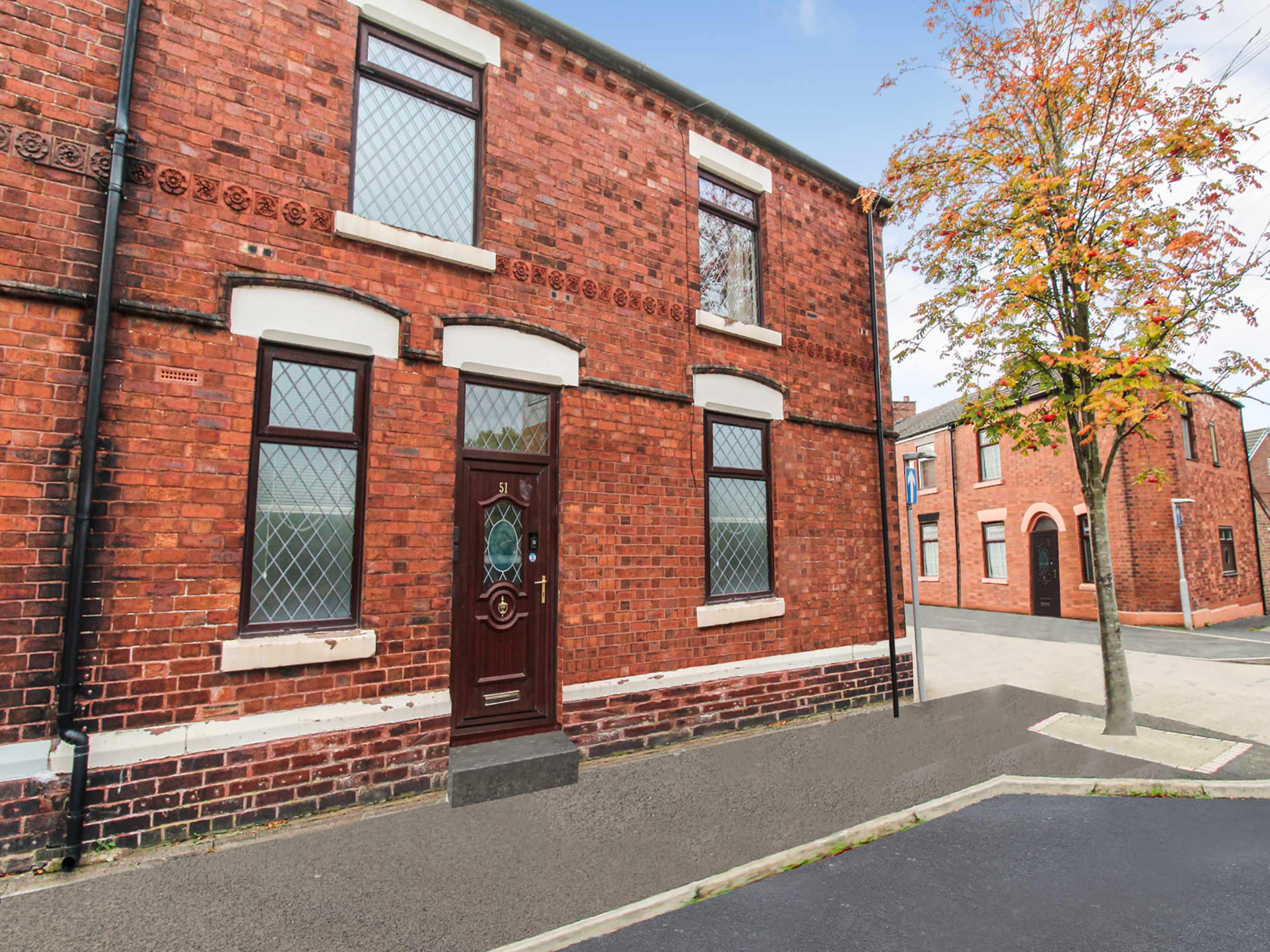 A red brick building with a decorative front door and diamond-patterned windows, situated at the corner of a street lined with similar structures and a tree.