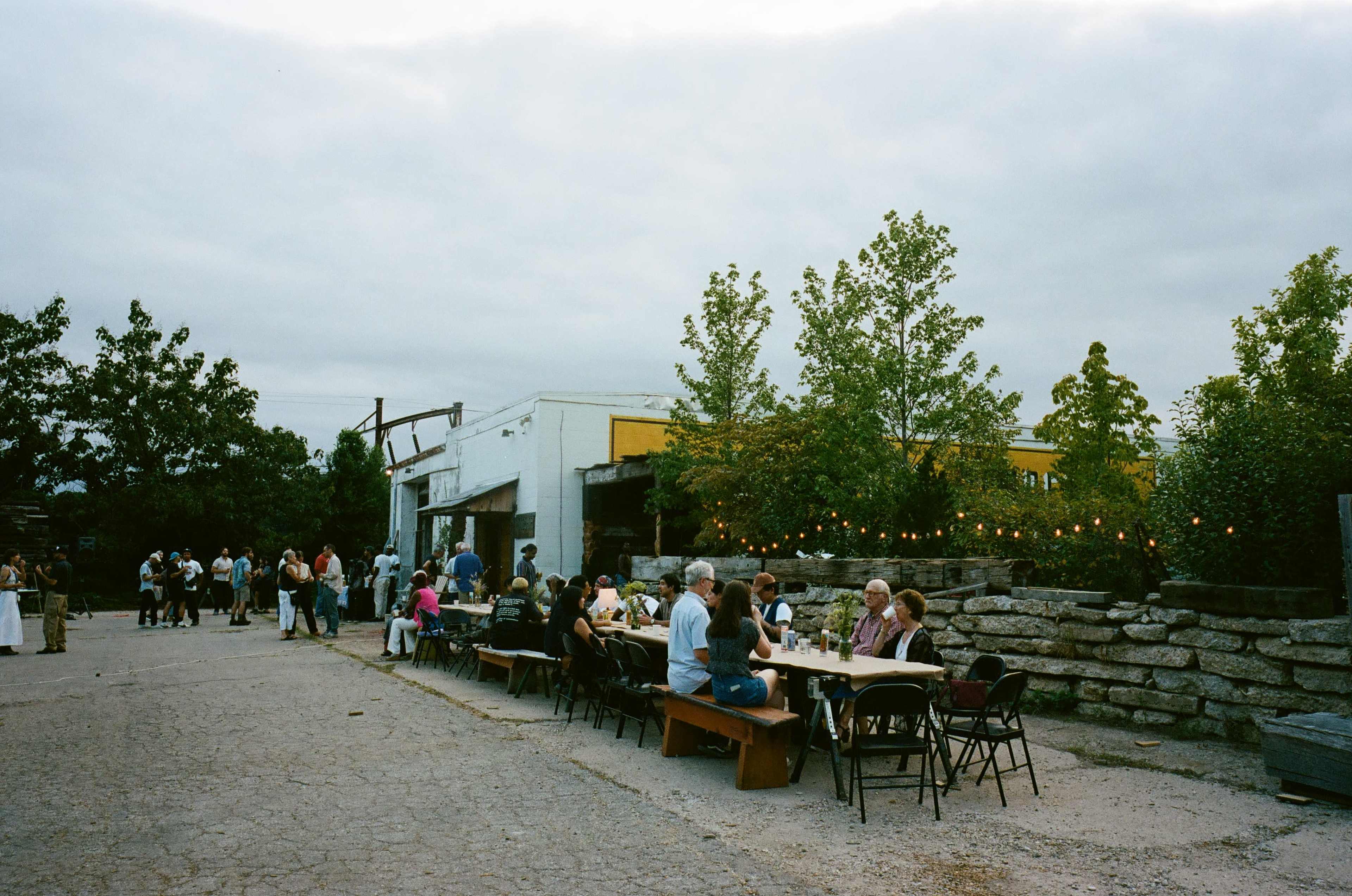 The image shows a gathering of people seated at tables and mingling in an outdoor setting, surrounded by trees and buildings under a cloudy sky.