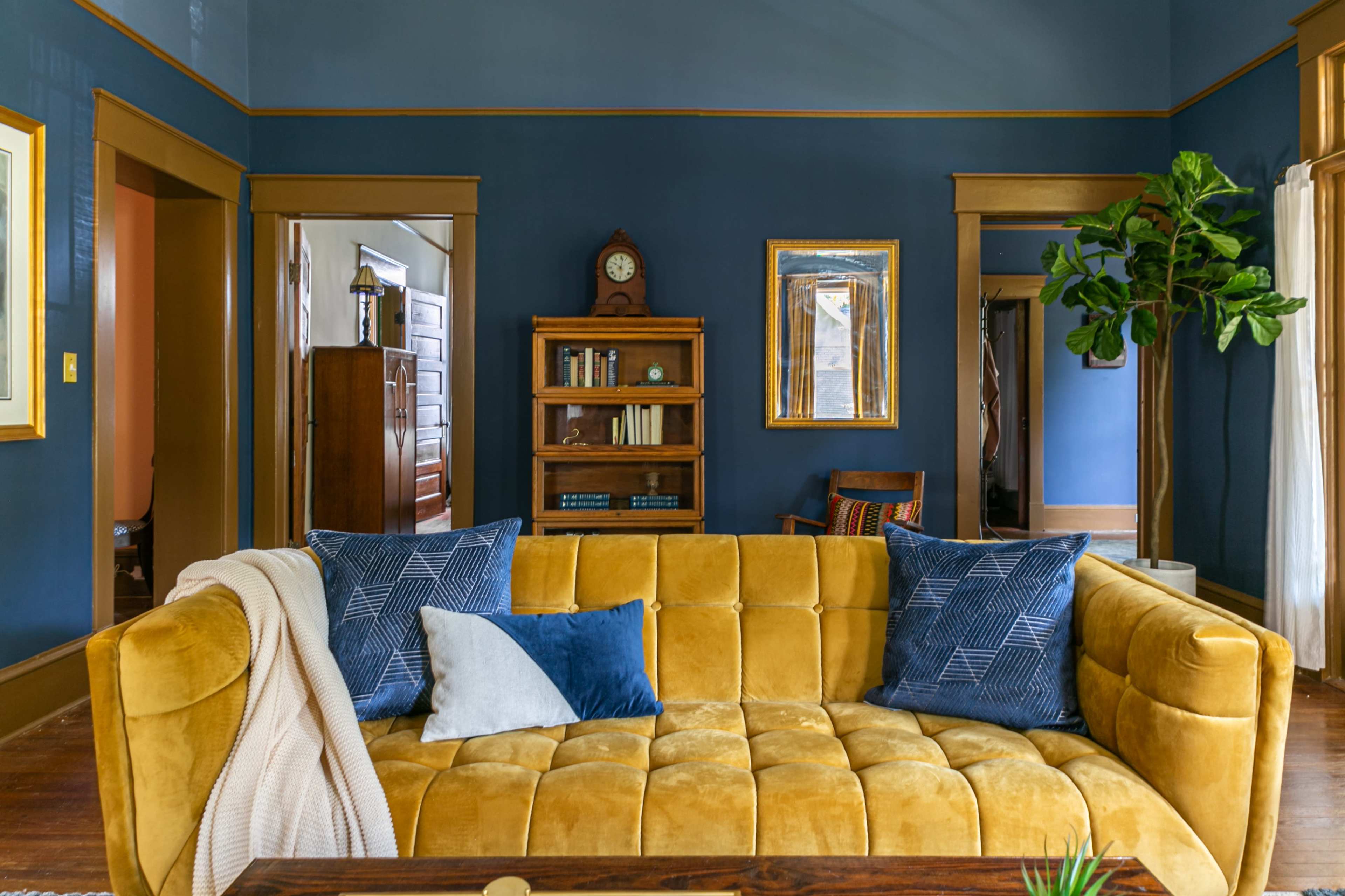 A cozy living room with a yellow tufted sofa, blue accent pillows, and walls painted in a dark shade, complemented by wooden accents and a decorative bookshelf.