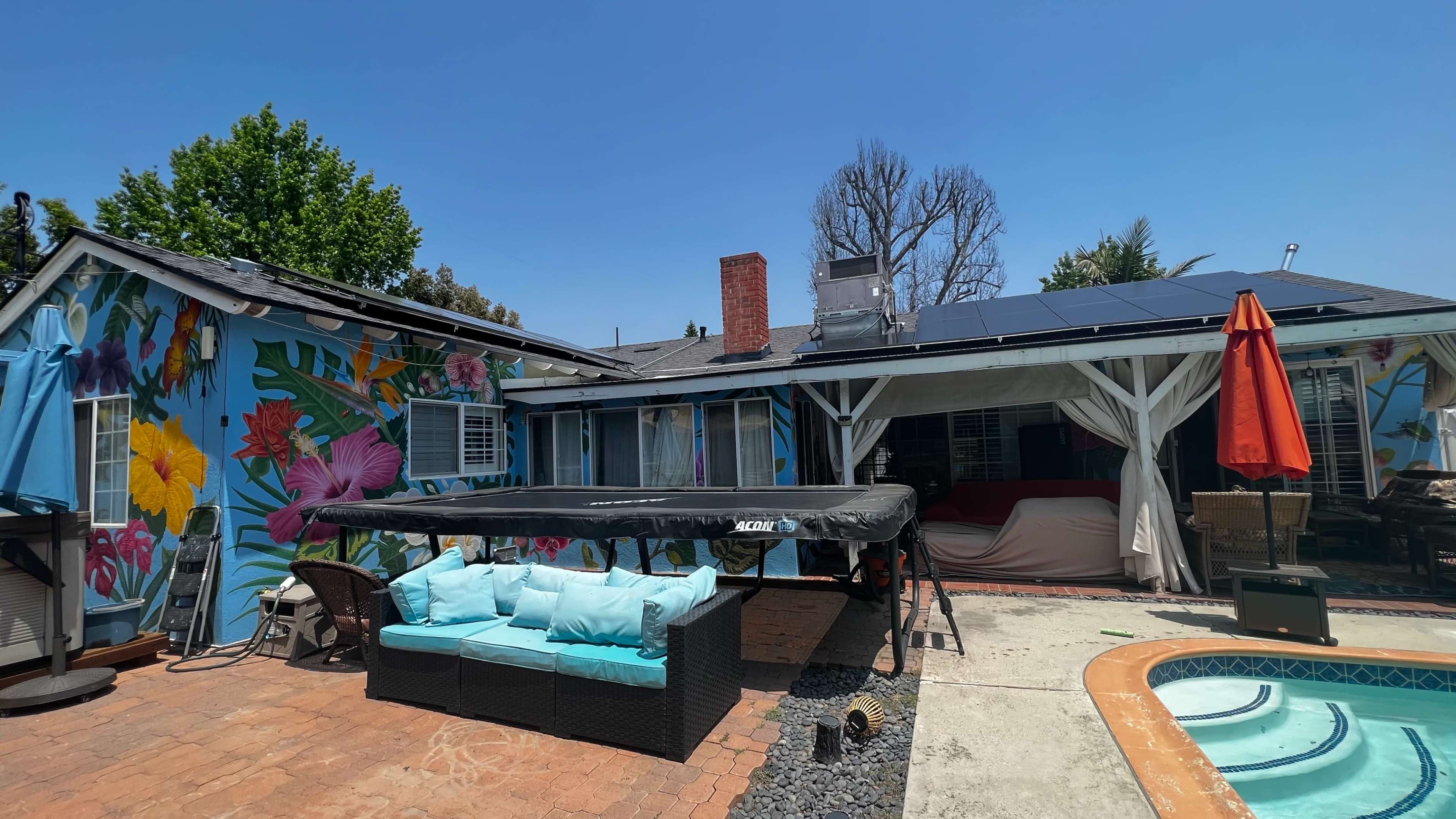 A colorful house with a mural, a patio area featuring a black wicker sofa with blue cushions, a solar panel roof, and a swimming pool nearby.