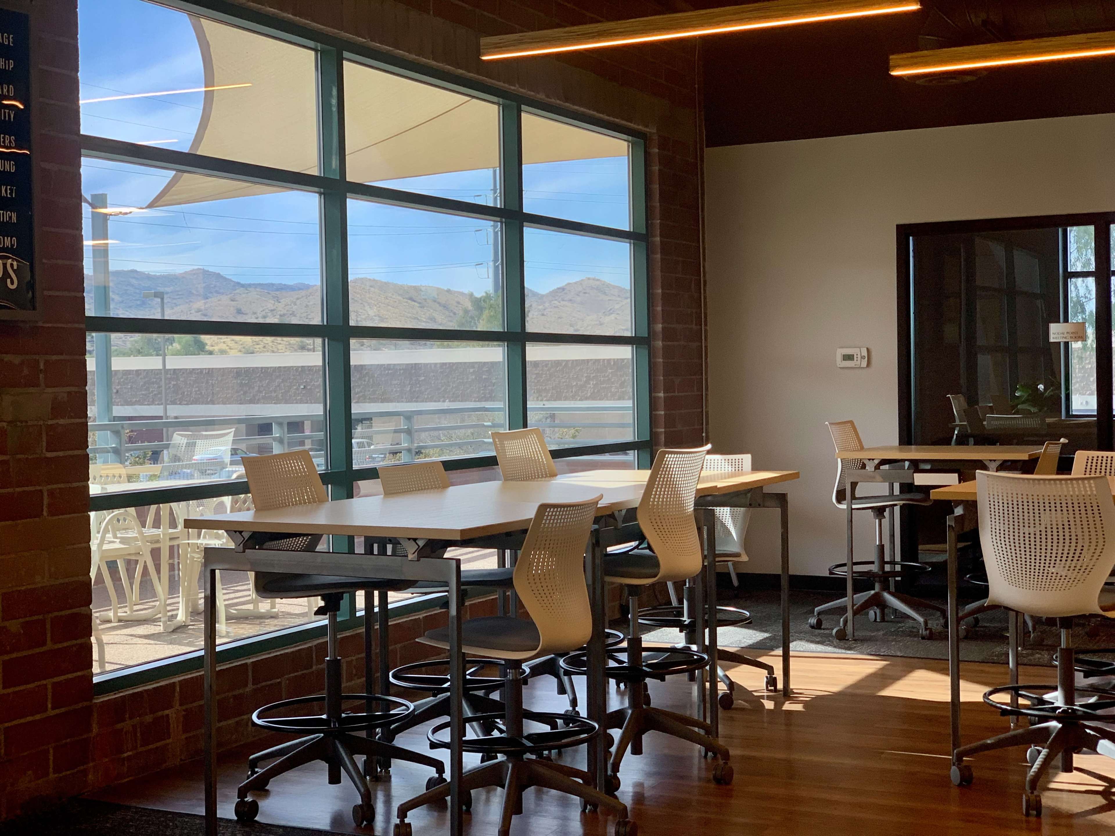 A bright, modern workspace featuring several high tables with stools beside large windows that overlook a scenic outdoor area with mountains in the background.