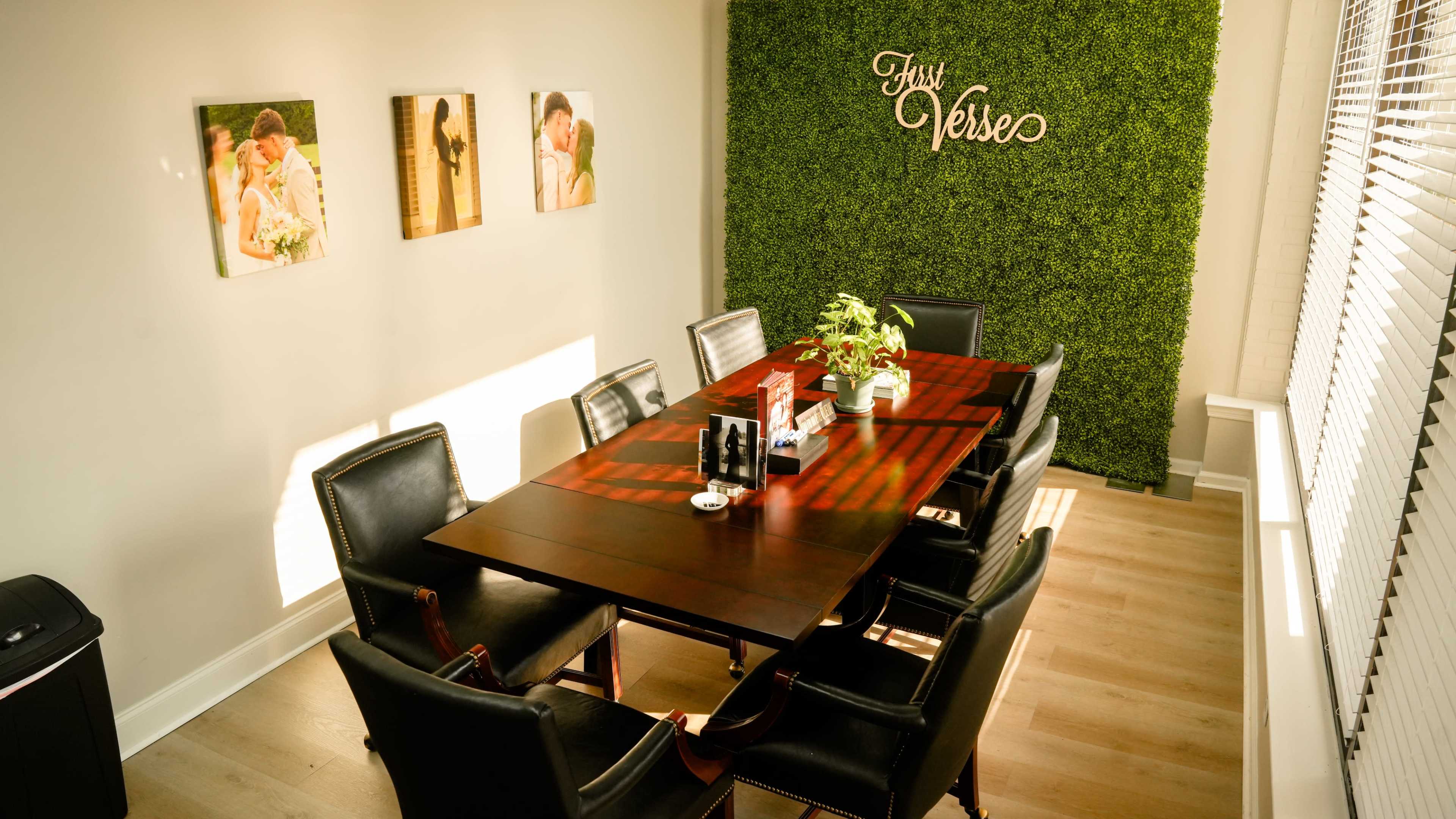 A conference room features a large wooden table surrounded by black chairs, with a green wall and framed photographs on the adjacent wall.