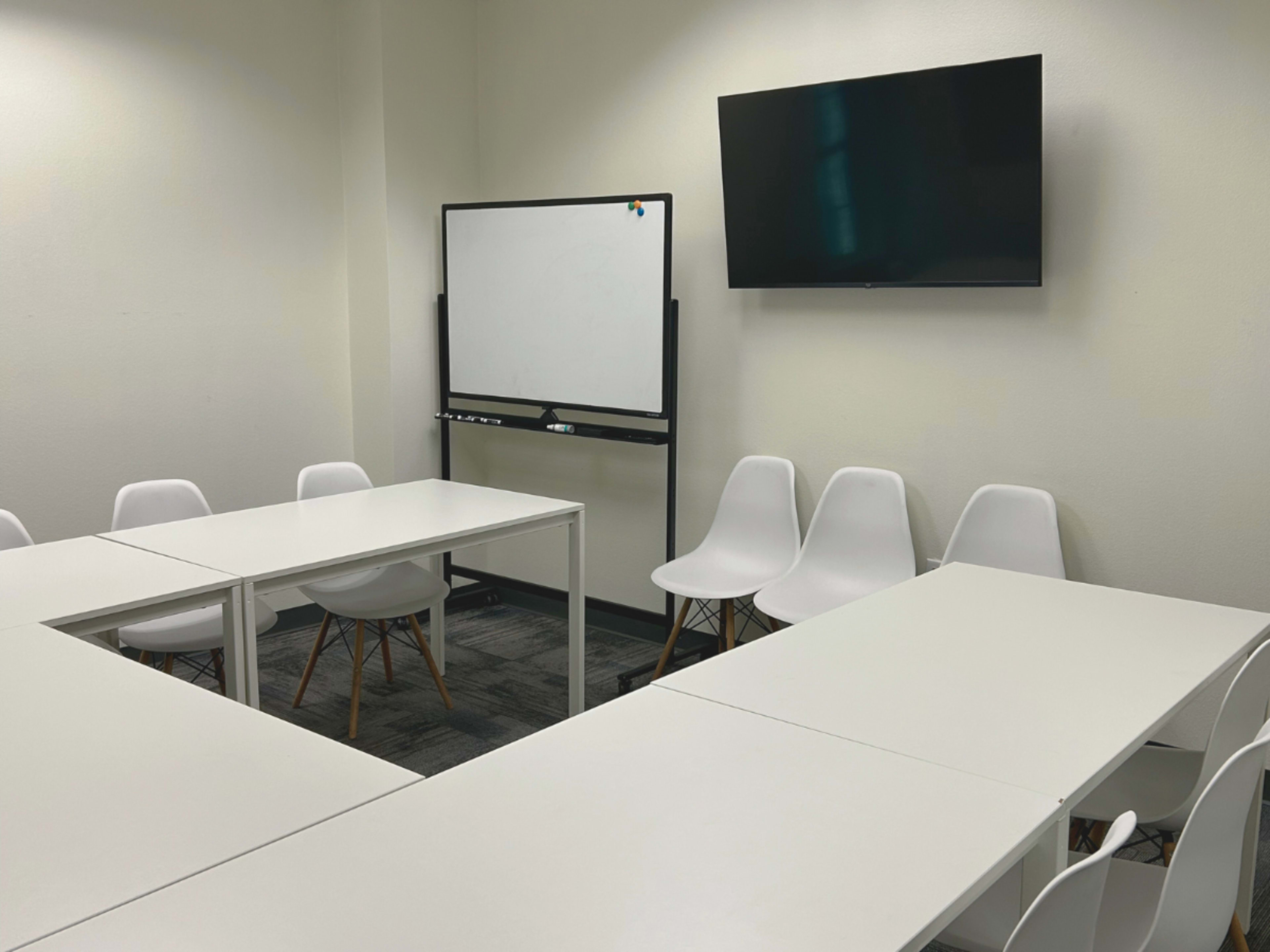The image shows a blank meeting room with a whiteboard on a stand, a wall-mounted television, and several white tables and chairs arranged around the space.