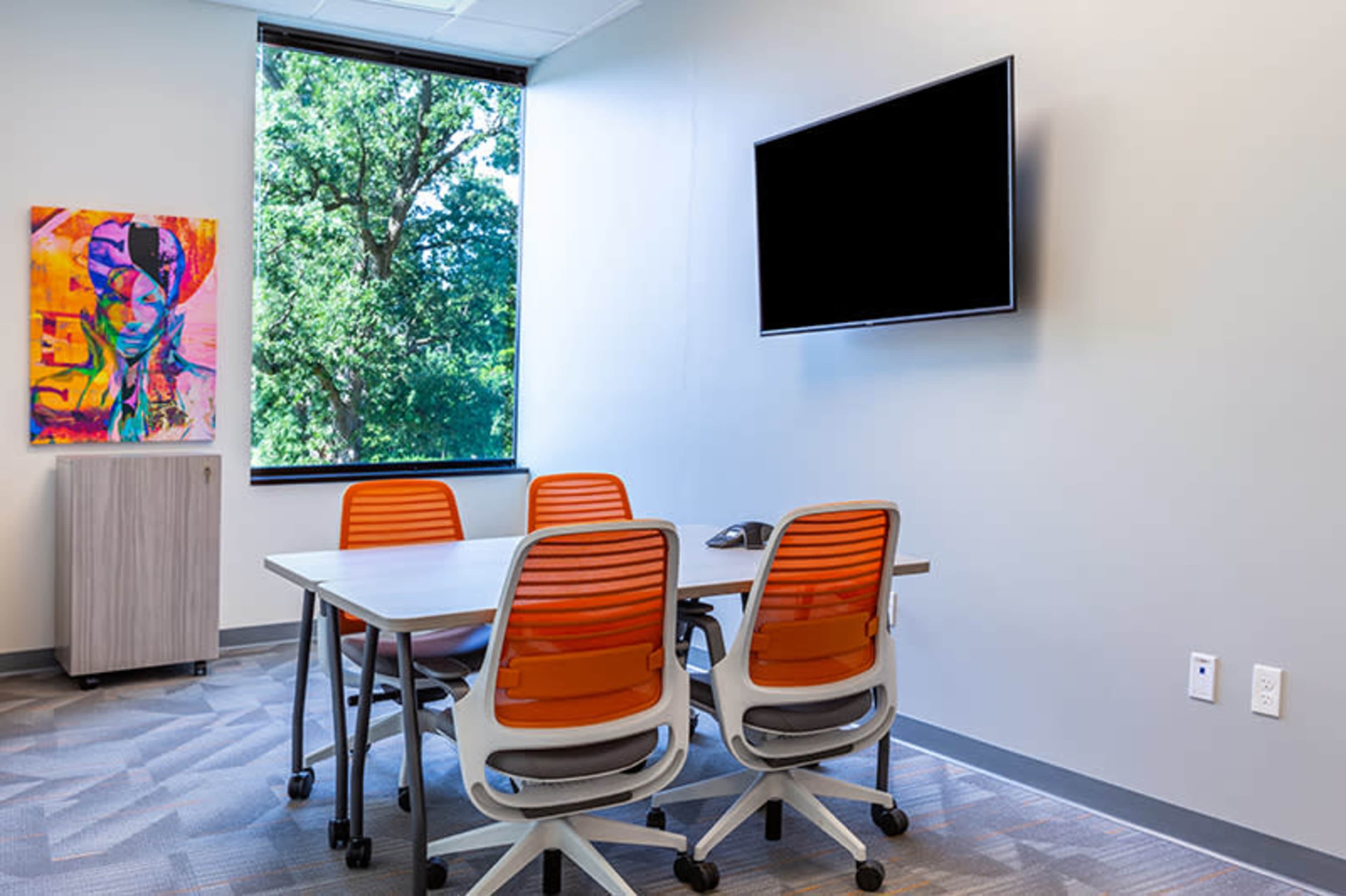 A conference room features a table with four orange chairs, a wall-mounted television, and a window displaying greenery outside.