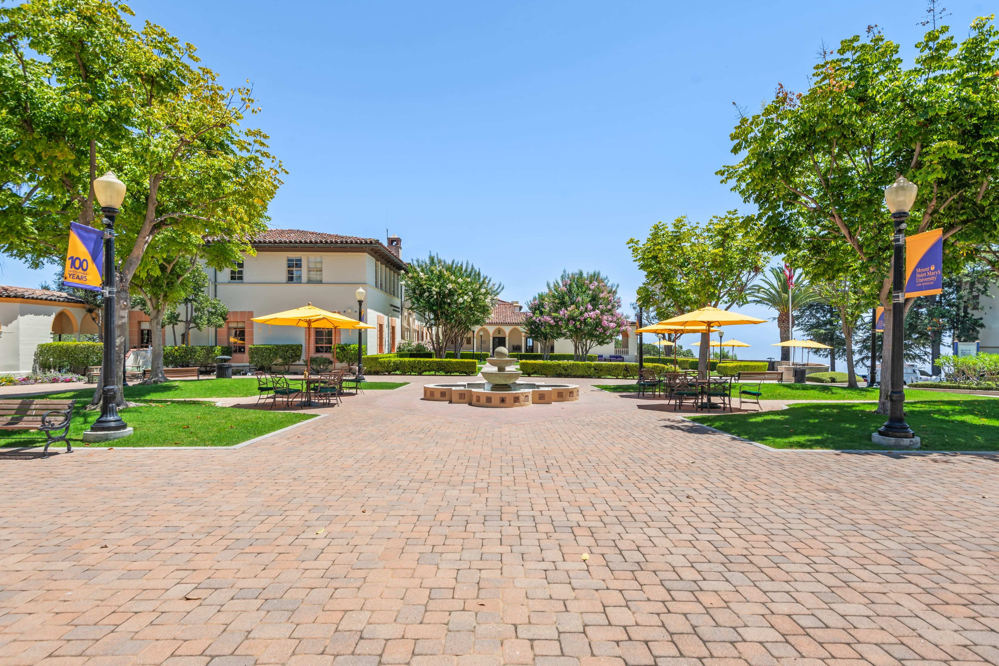 The image shows a landscaped courtyard with a fountain at the center, surrounded by tables, chairs, and trees, with buildings visible in the background.