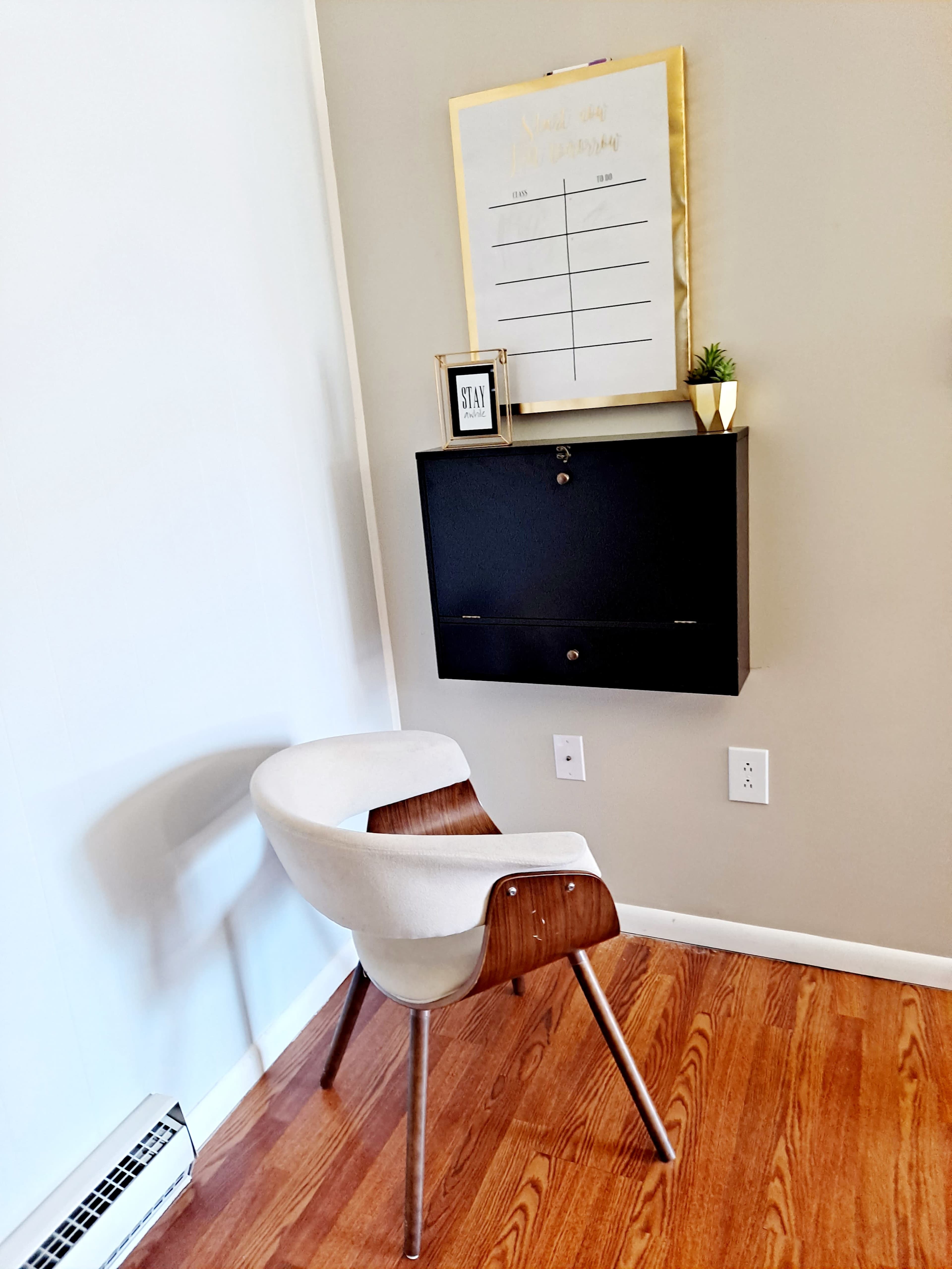 A white chair with wooden accents sits beside a wall-mounted black cabinet and a framed schedule on the wall.