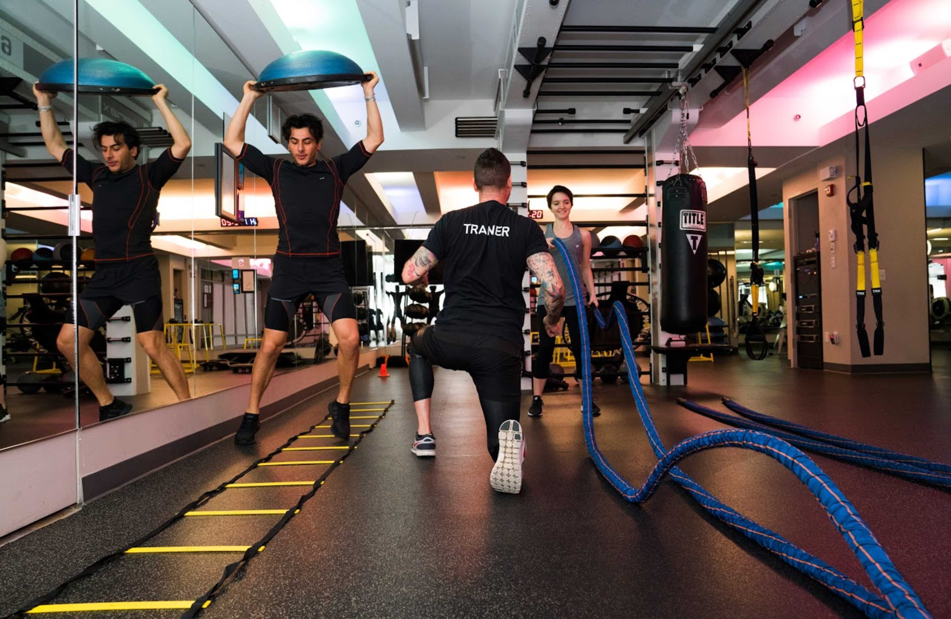 A trainer supervises a client performing an exercise with a stability ball in a gym equipped with various fitness equipment.