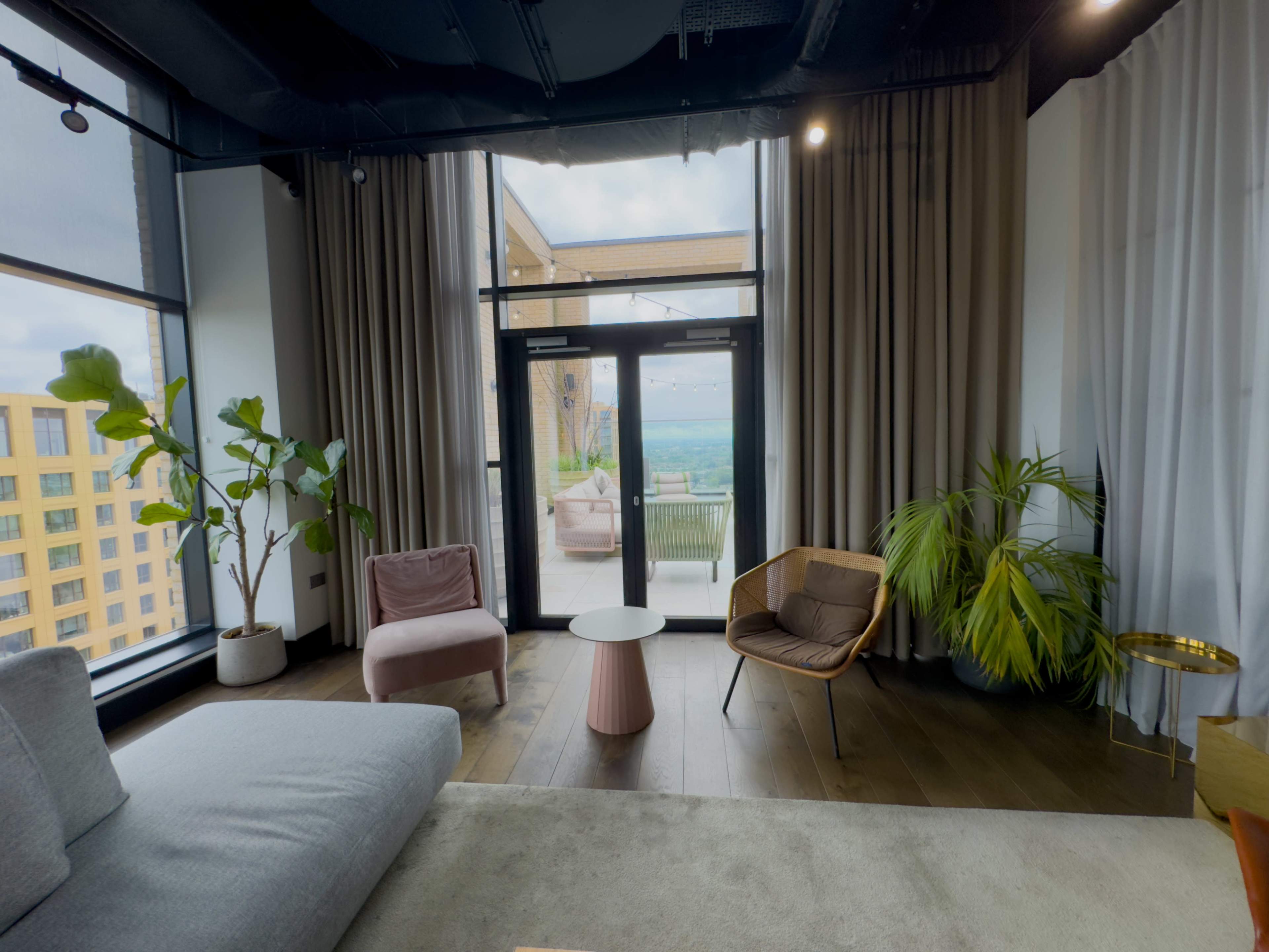 The image shows a modern living room with large windows, a gray sofa, two armchairs, a small round table, and indoor plants.