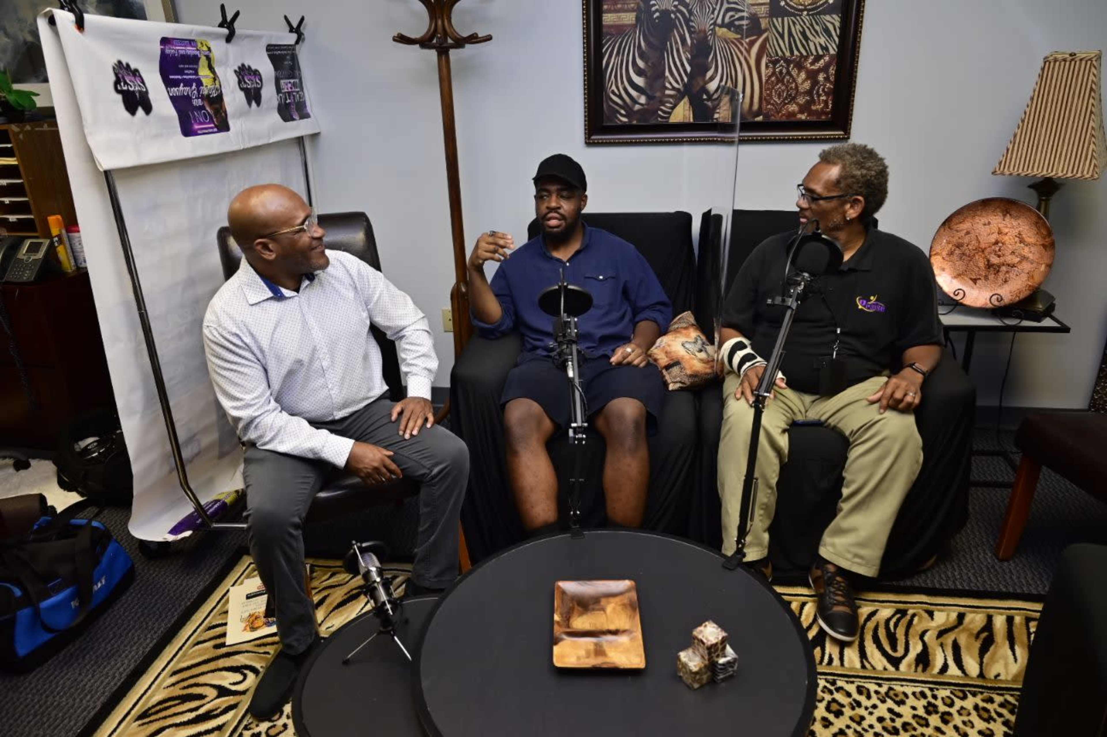 Three men are seated in a room with a zebra-print rug, engaging in conversation next to a small round table.