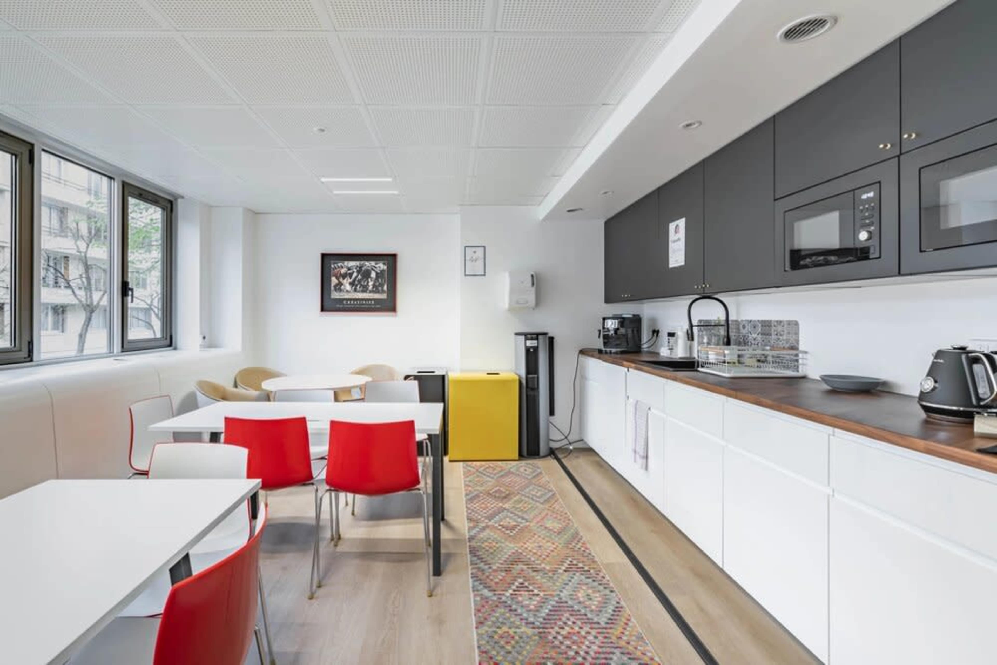 The image shows a modern kitchen and dining area featuring a mixed arrangement of white tables and red chairs, equipped with appliances and a colorful yellow cabinet.