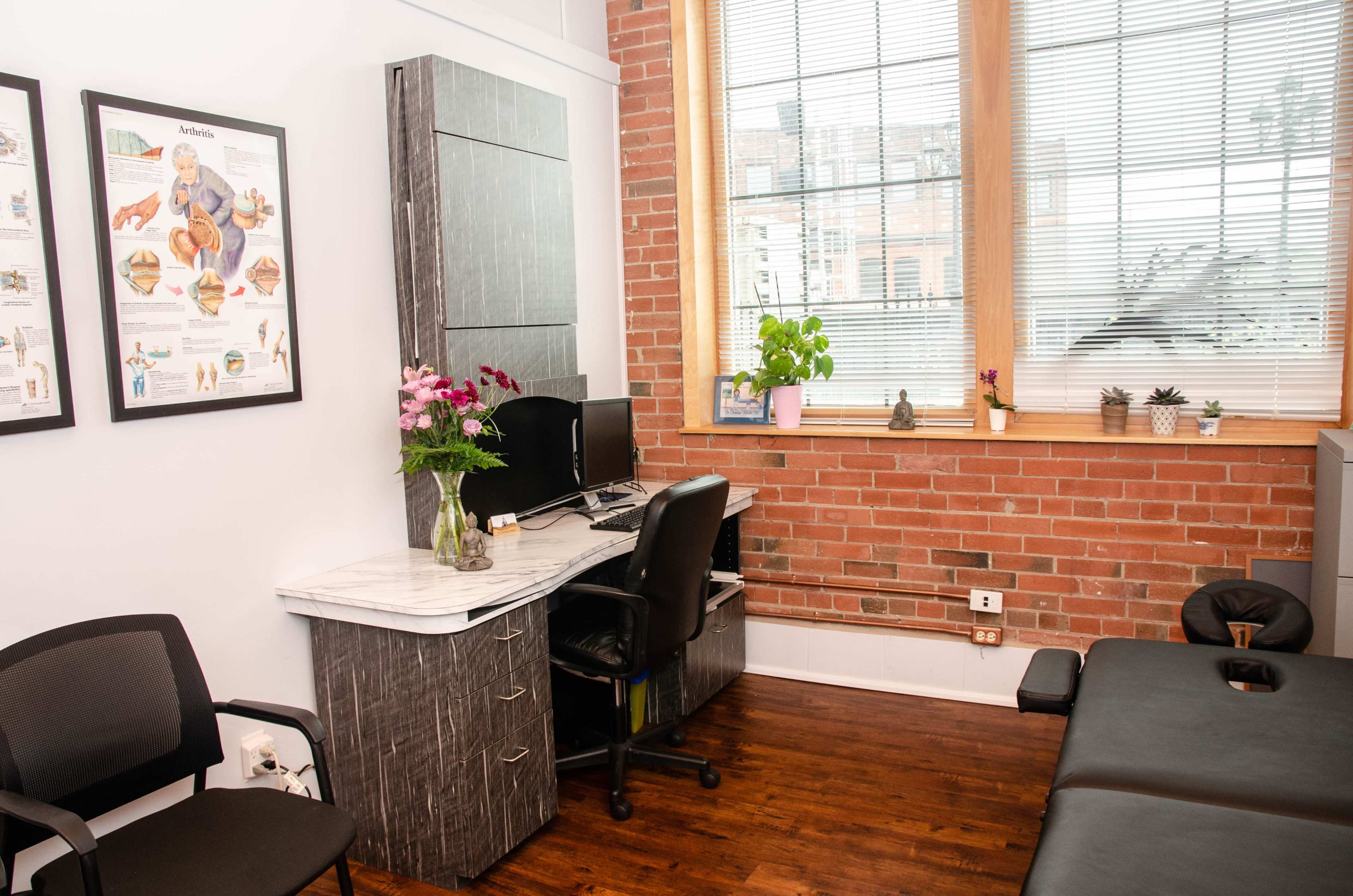 A therapy office featuring a desk with a computer, a chair, and a treatment table, all set against a backdrop of exposed brick walls and large windows.