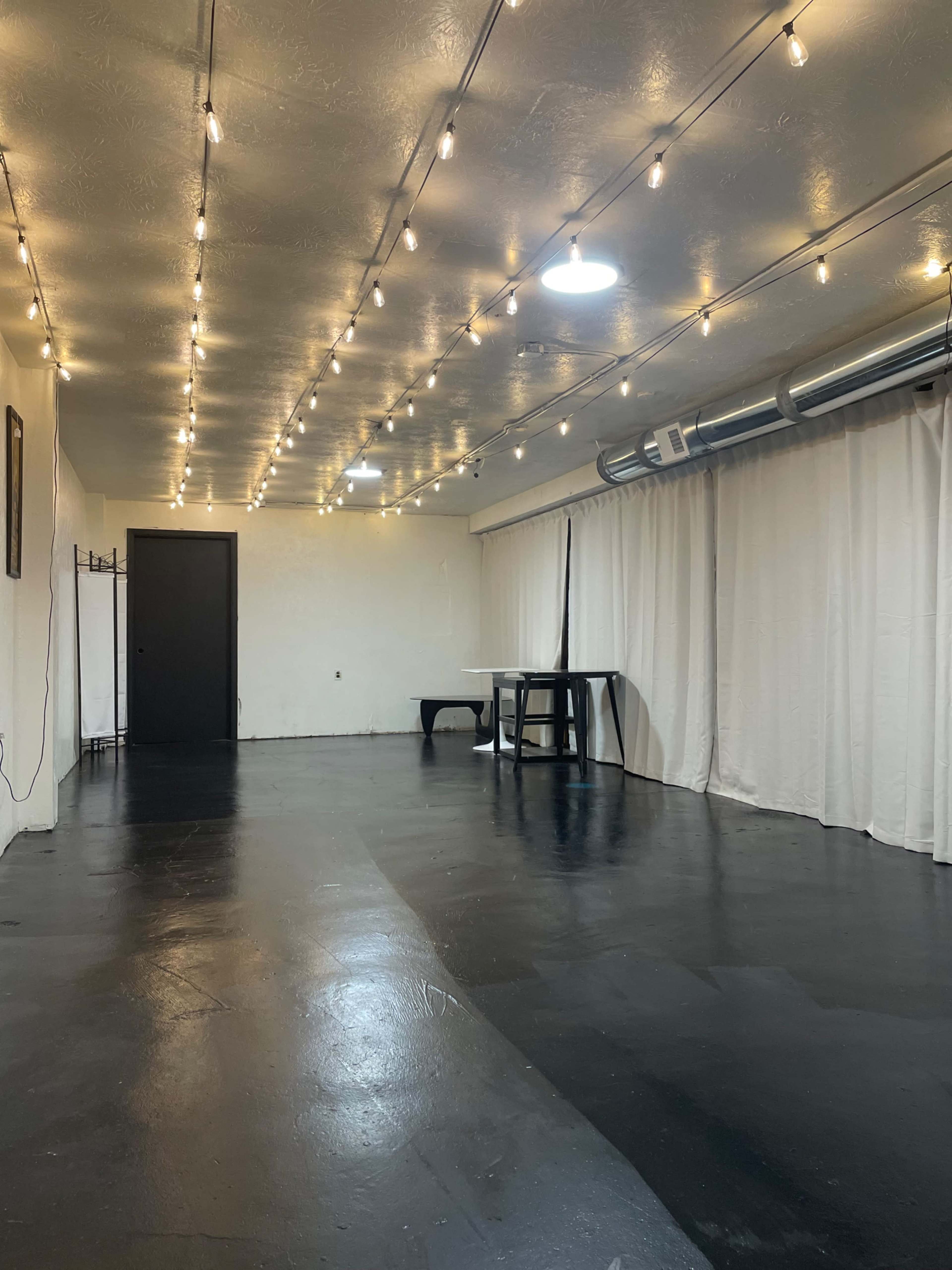 The image shows an empty room with black floors, white curtains, and string lights overhead.