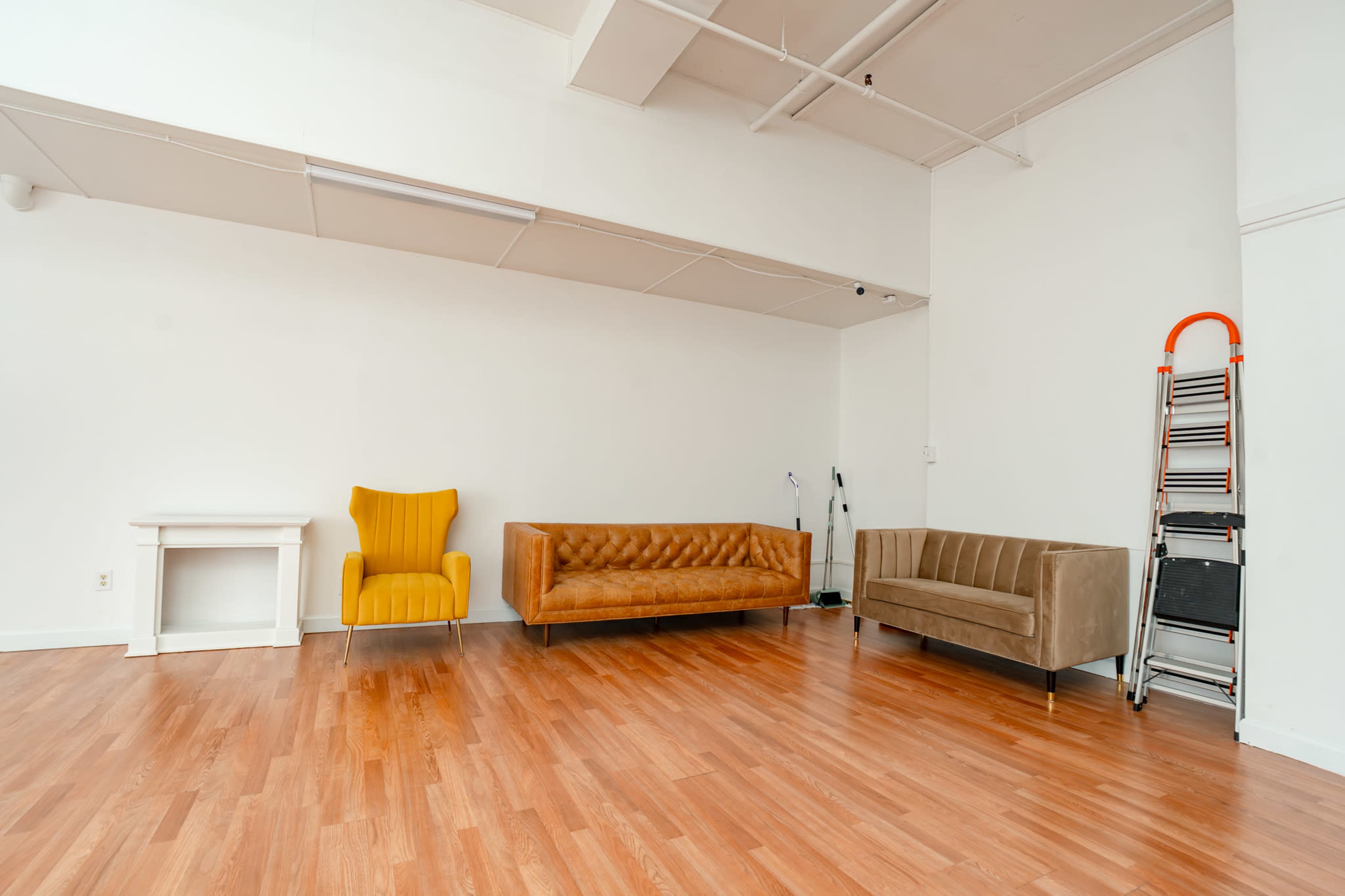 The image shows a spacious, empty room with wooden flooring, featuring a yellow chair, a brown sofa, and a gray sofa, along with a ladder against the wall.