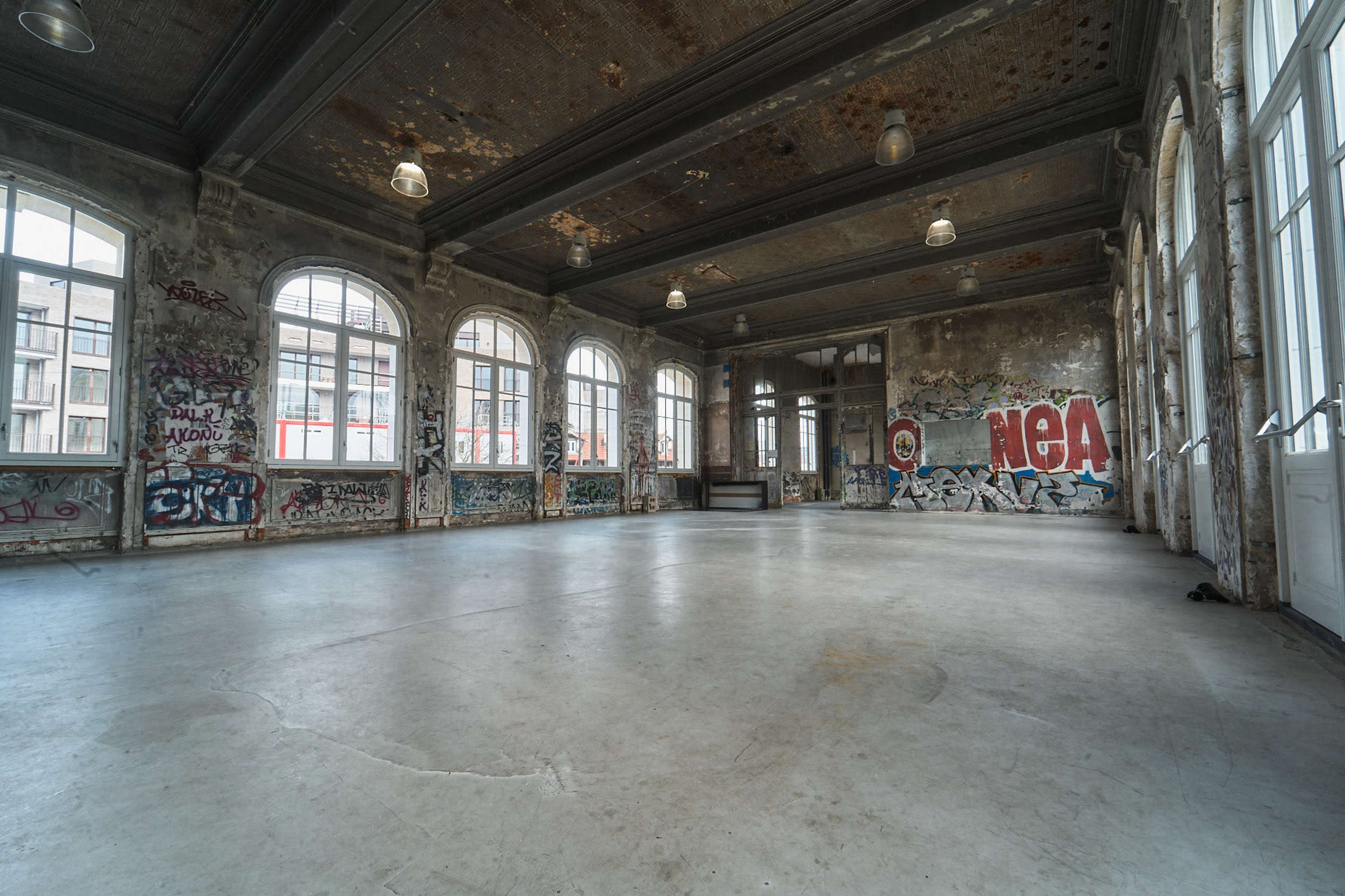 The image shows an empty, dilapidated room with large windows, exposed brick walls, and graffiti covering the surfaces.