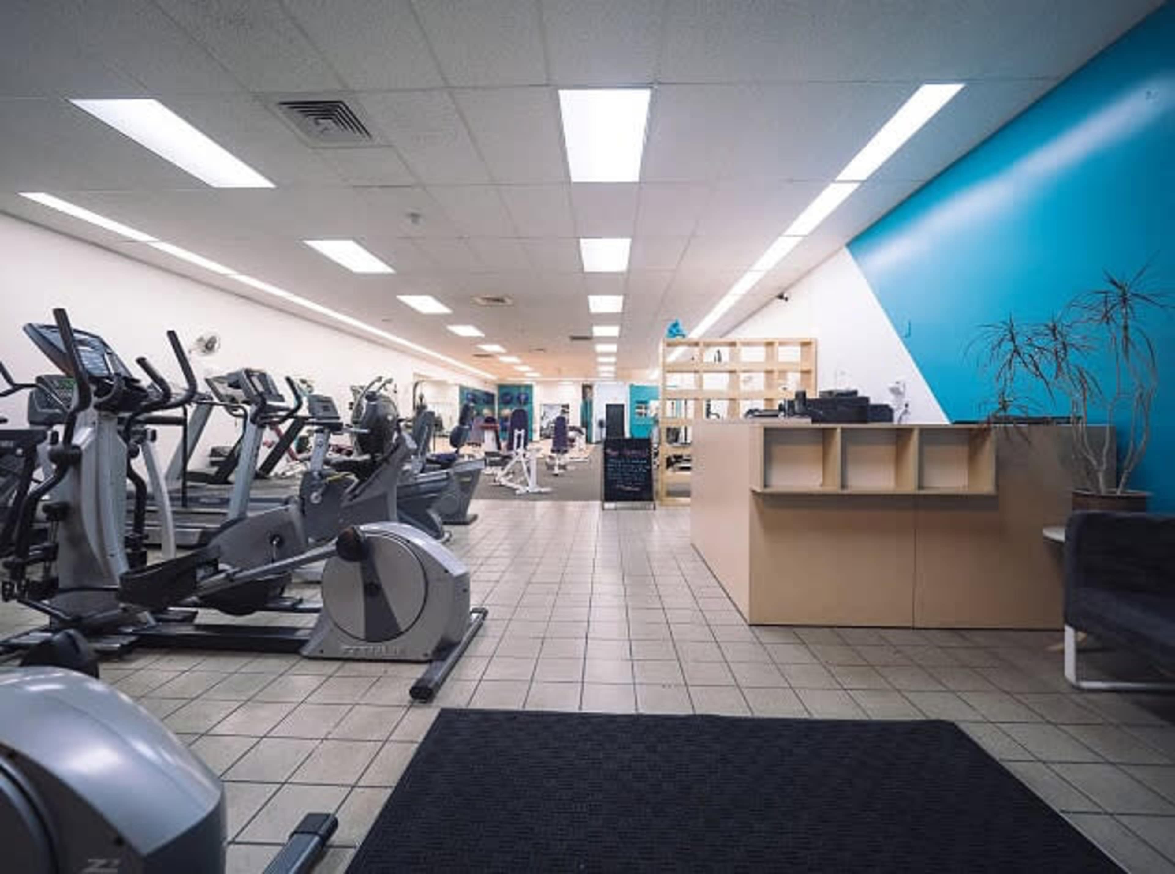 A gym interior featuring exercise equipment arranged along one side, with a reception area visible in the foreground.
