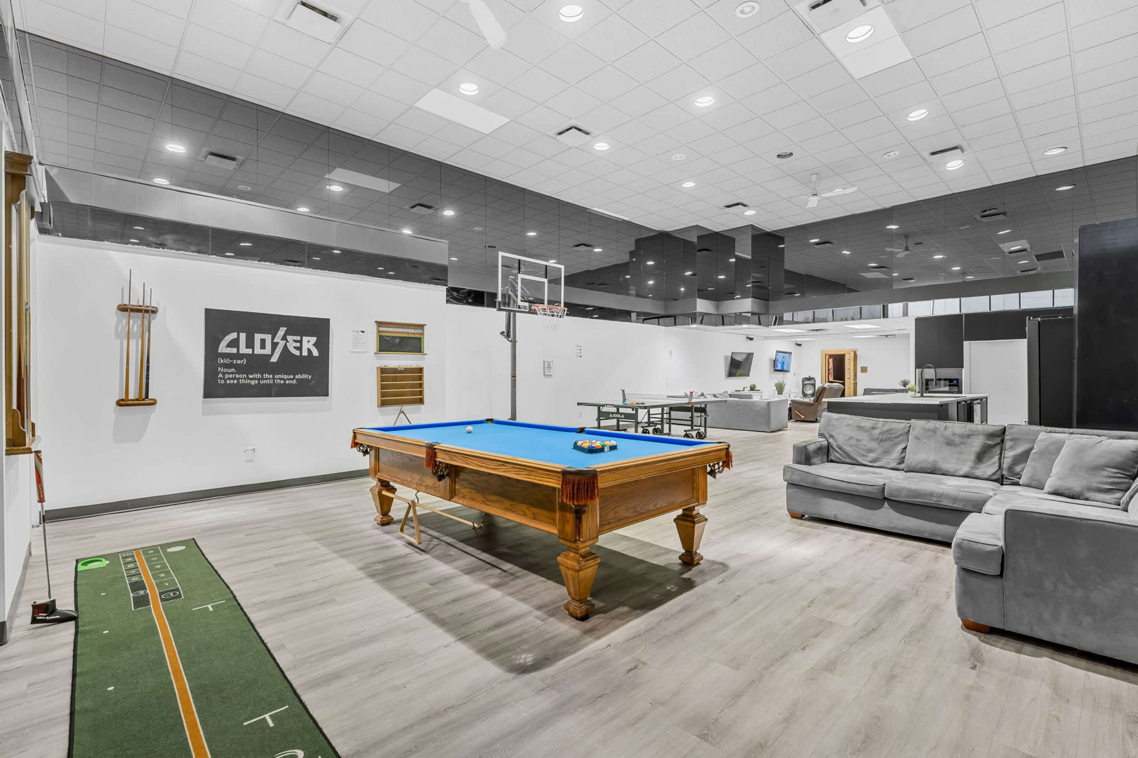 The image shows a game room featuring a pool table with blue felt, a gray sofa, a basketball hoop, and a green felt incline for a game.