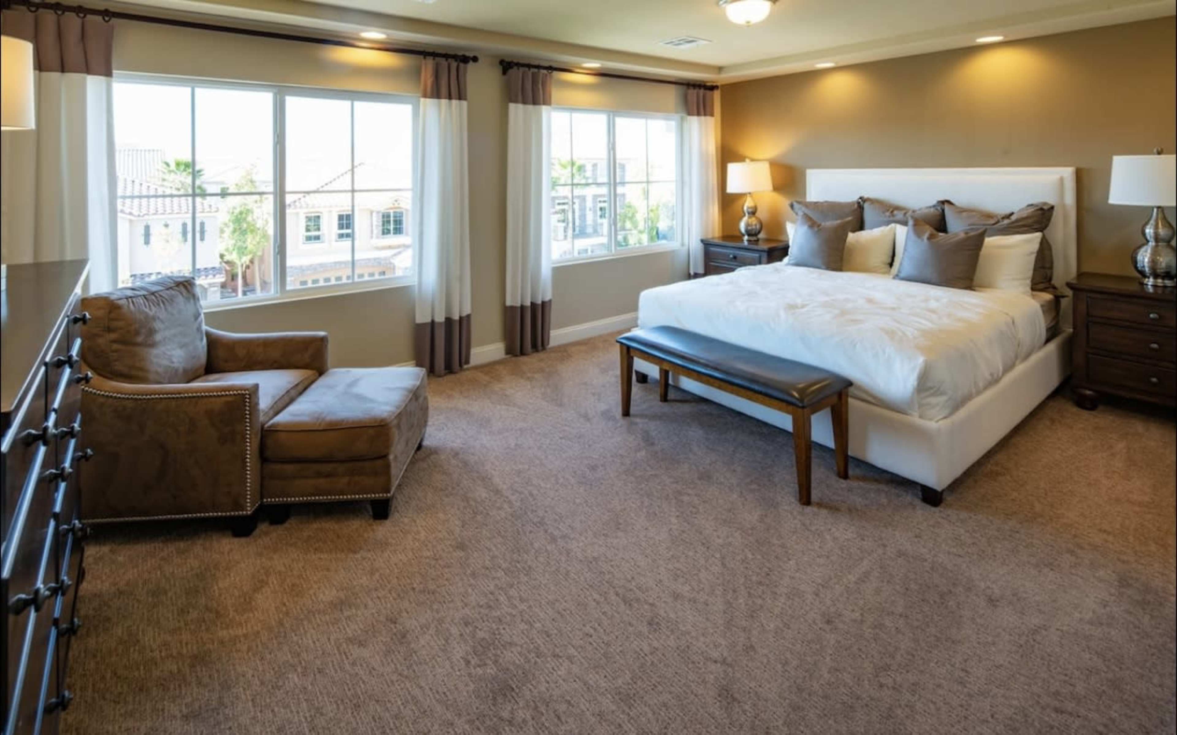 The image shows a well-furnished bedroom with a large bed, two bedside tables, a seating area with a chair, and large windows providing natural light.