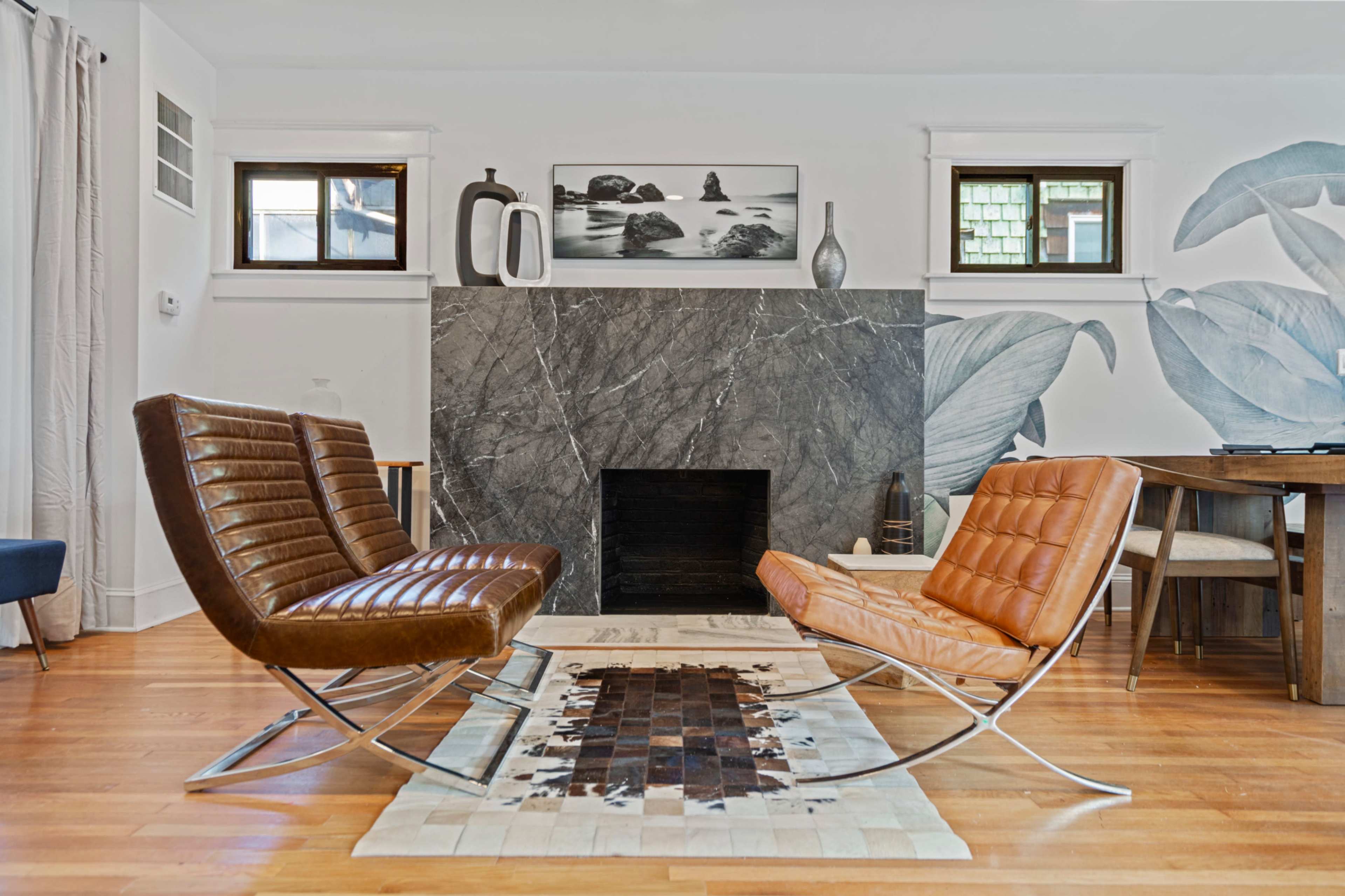 The living room features two modern chairs facing a marble fireplace, with a large wall mural of leaves and a wooden dining table nearby.