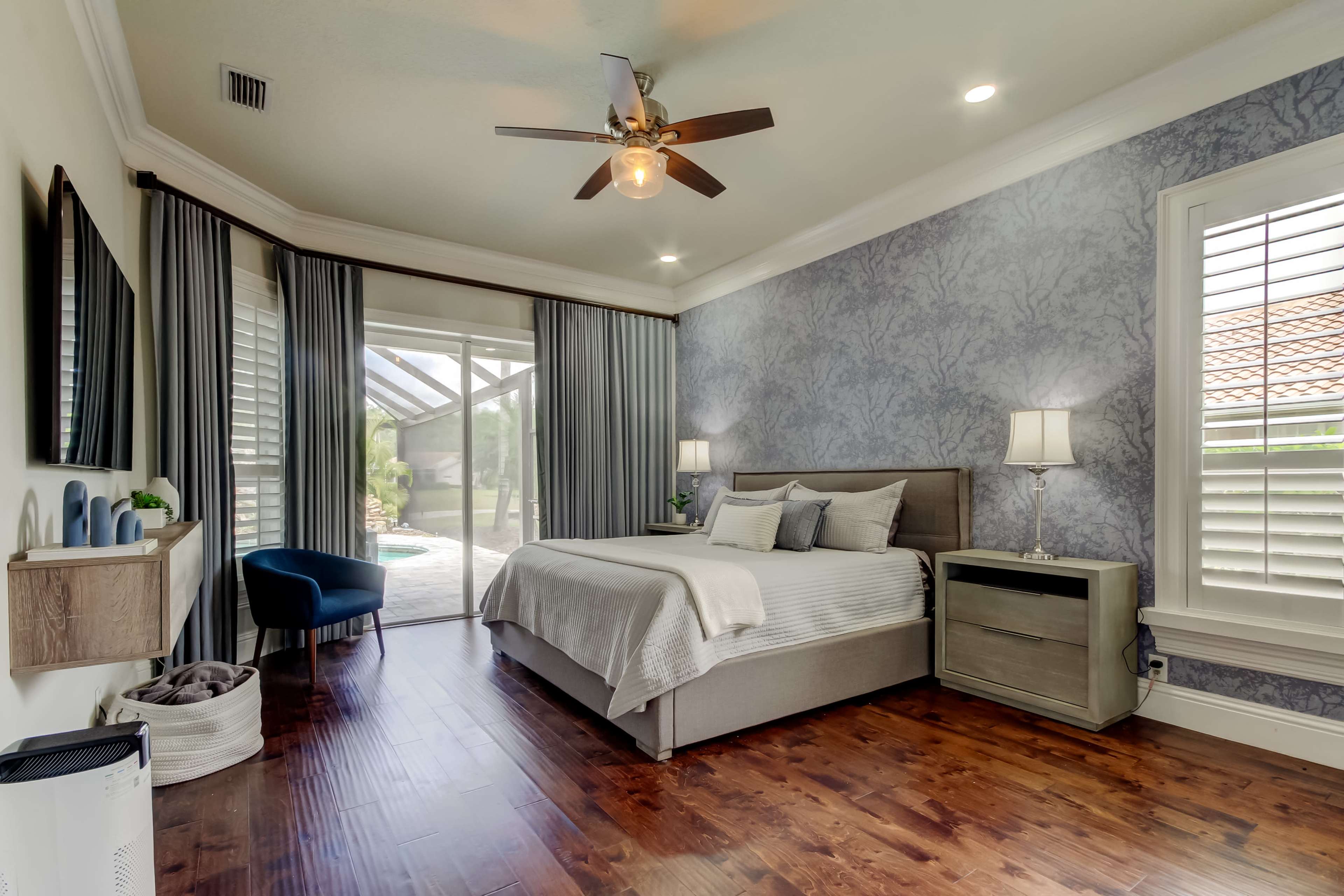 The image shows a modern bedroom featuring a bed with light bedding, a nightstand, a stylish chair, and large windows with sheer curtains allowing natural light to enter.
