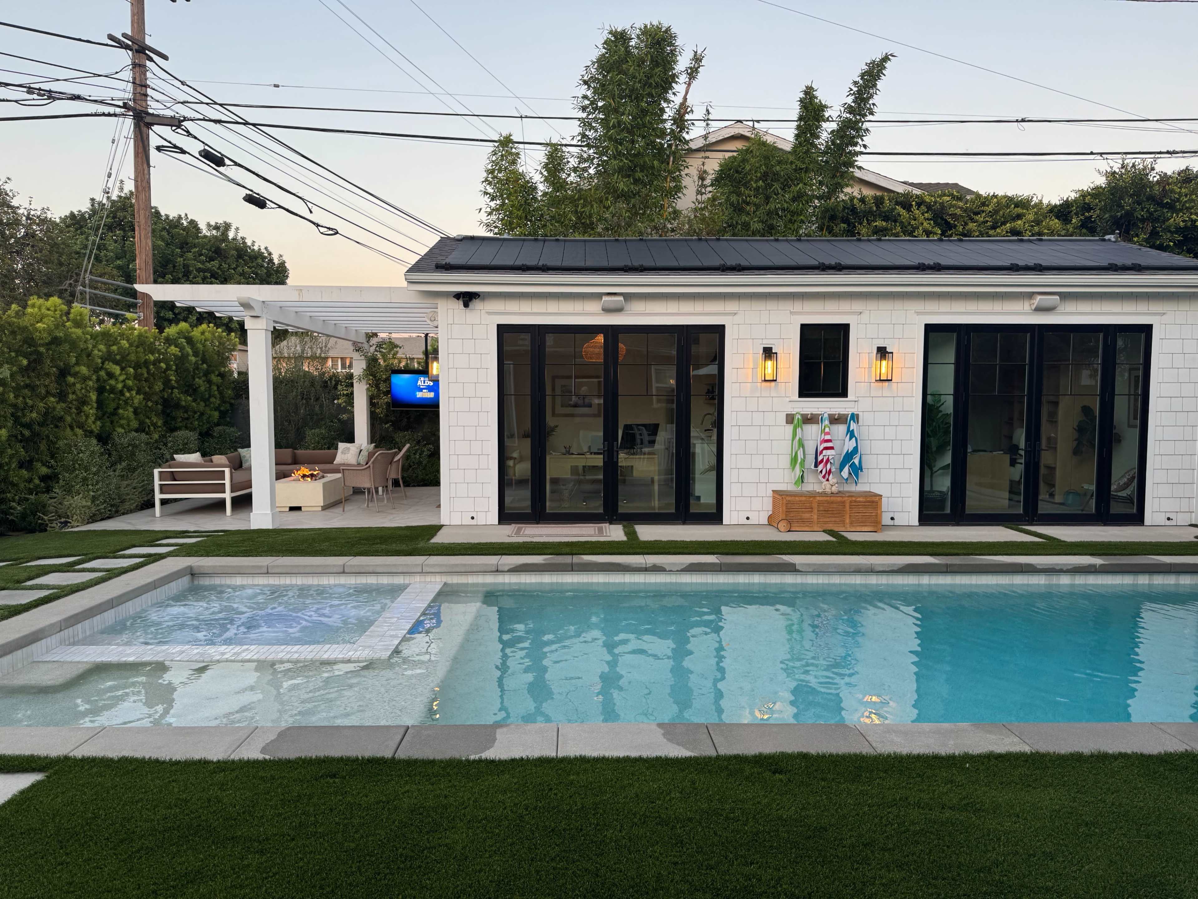 The image shows a modern backyard with a swimming pool, hot tub, and a white house featuring large black-framed windows and a patio area.