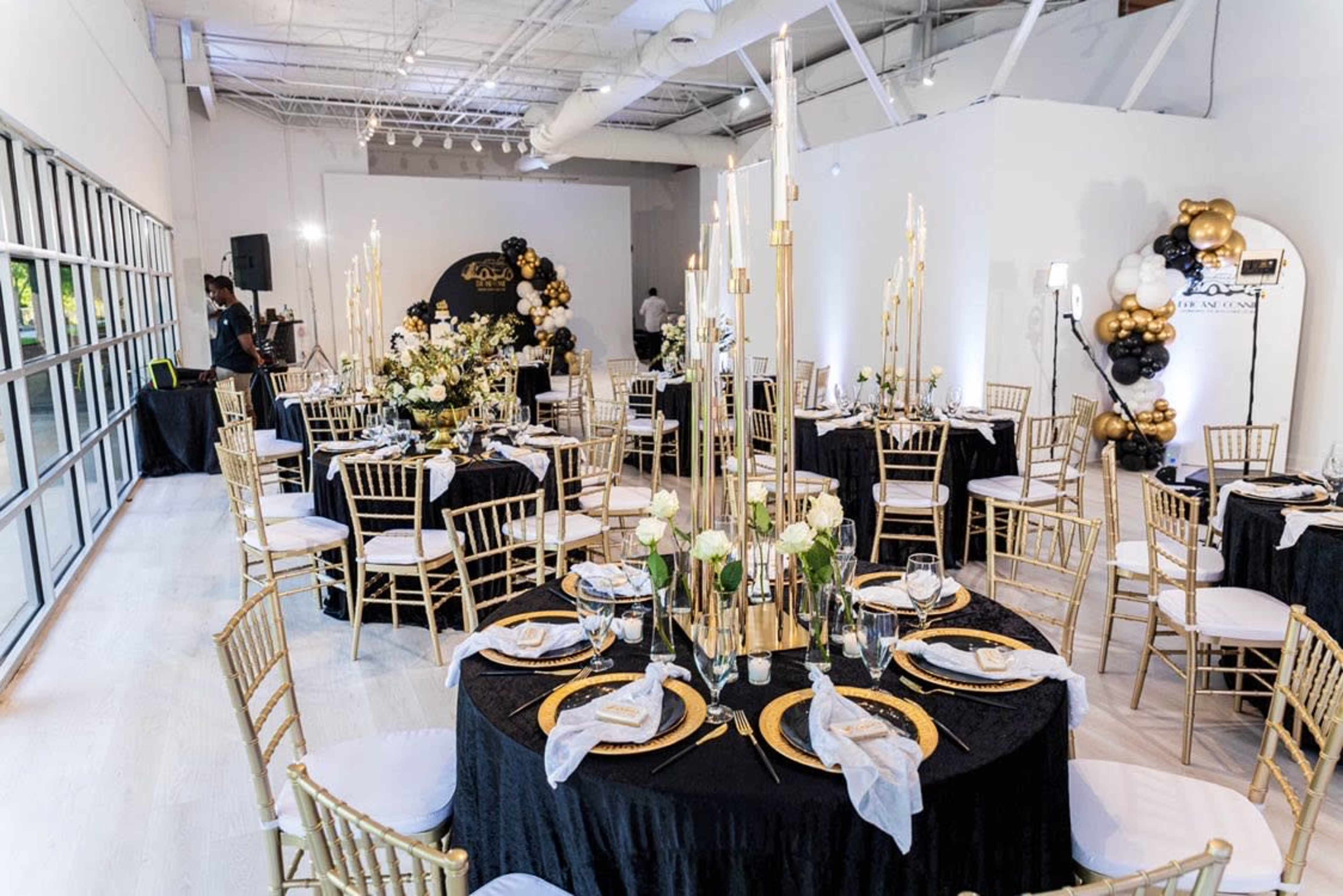 The image shows a banquet hall set up with black and gold table settings, featuring gold chiavari chairs, tall candles, and floral centerpieces.