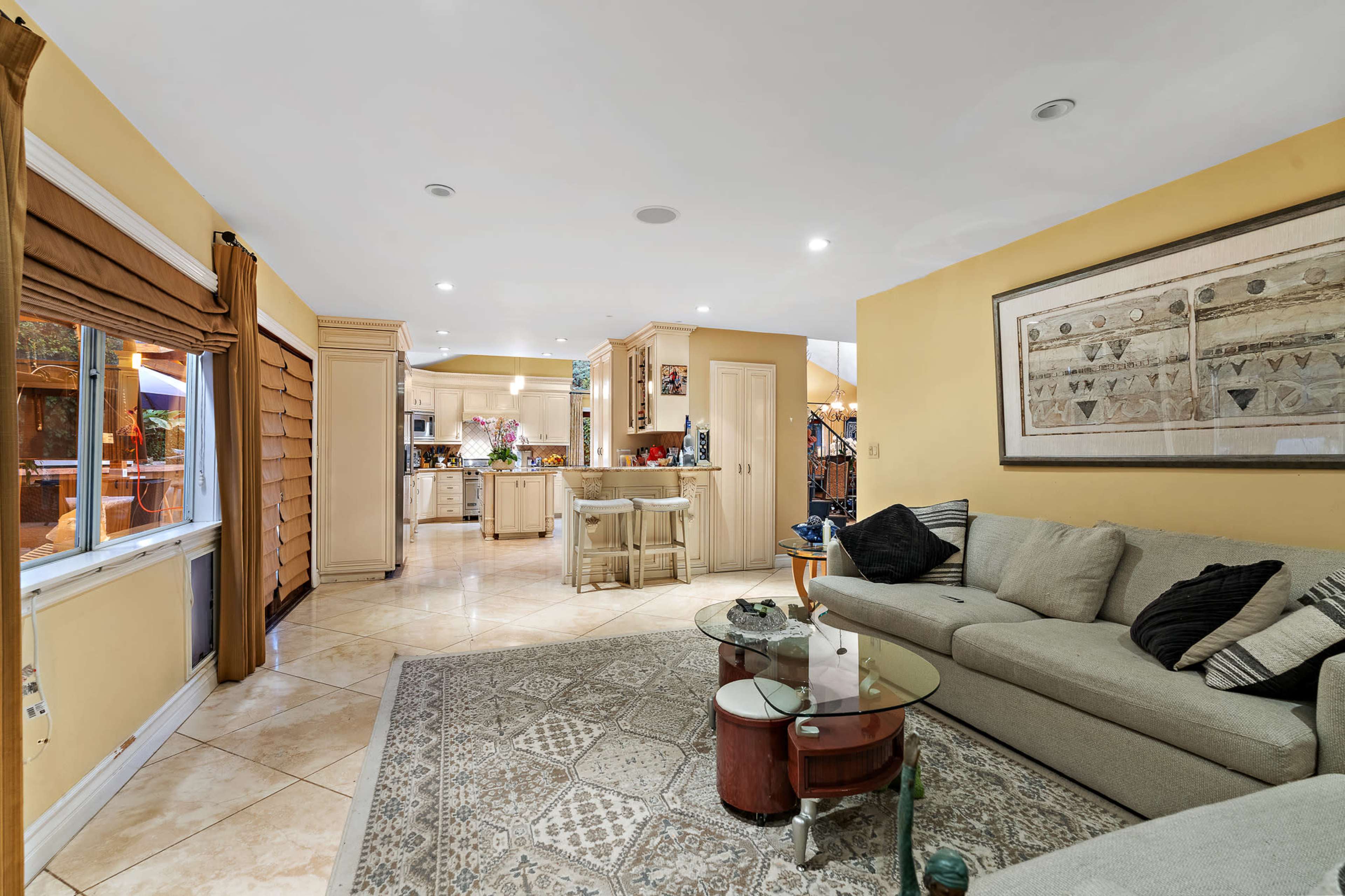 The image shows a spacious living room with a beige sectional sofa, a glass coffee table, and an open layout leading to a kitchen area.