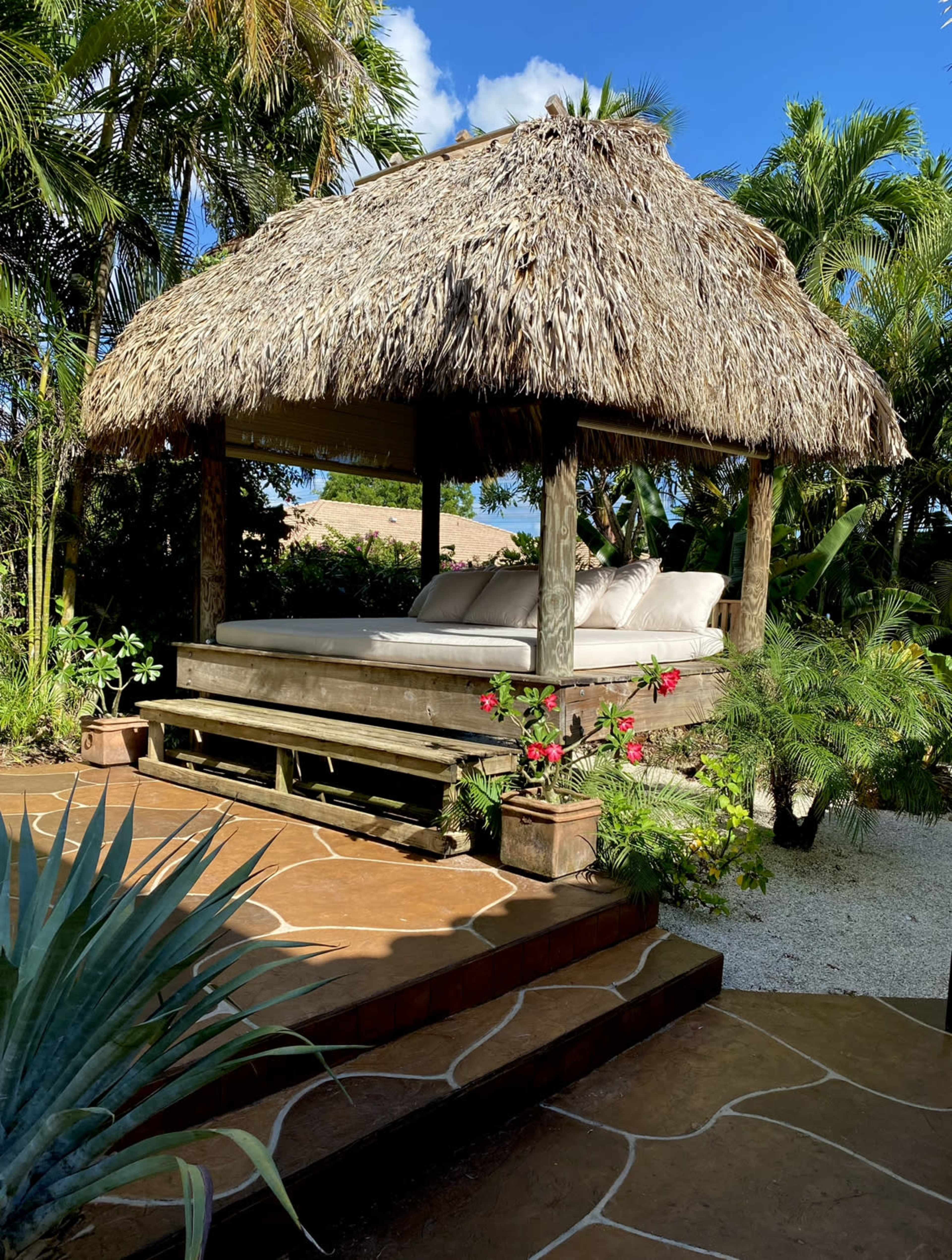 A thatched-roof gazebo with a cushioned seating area is set among tropical plants and stone pathways.
