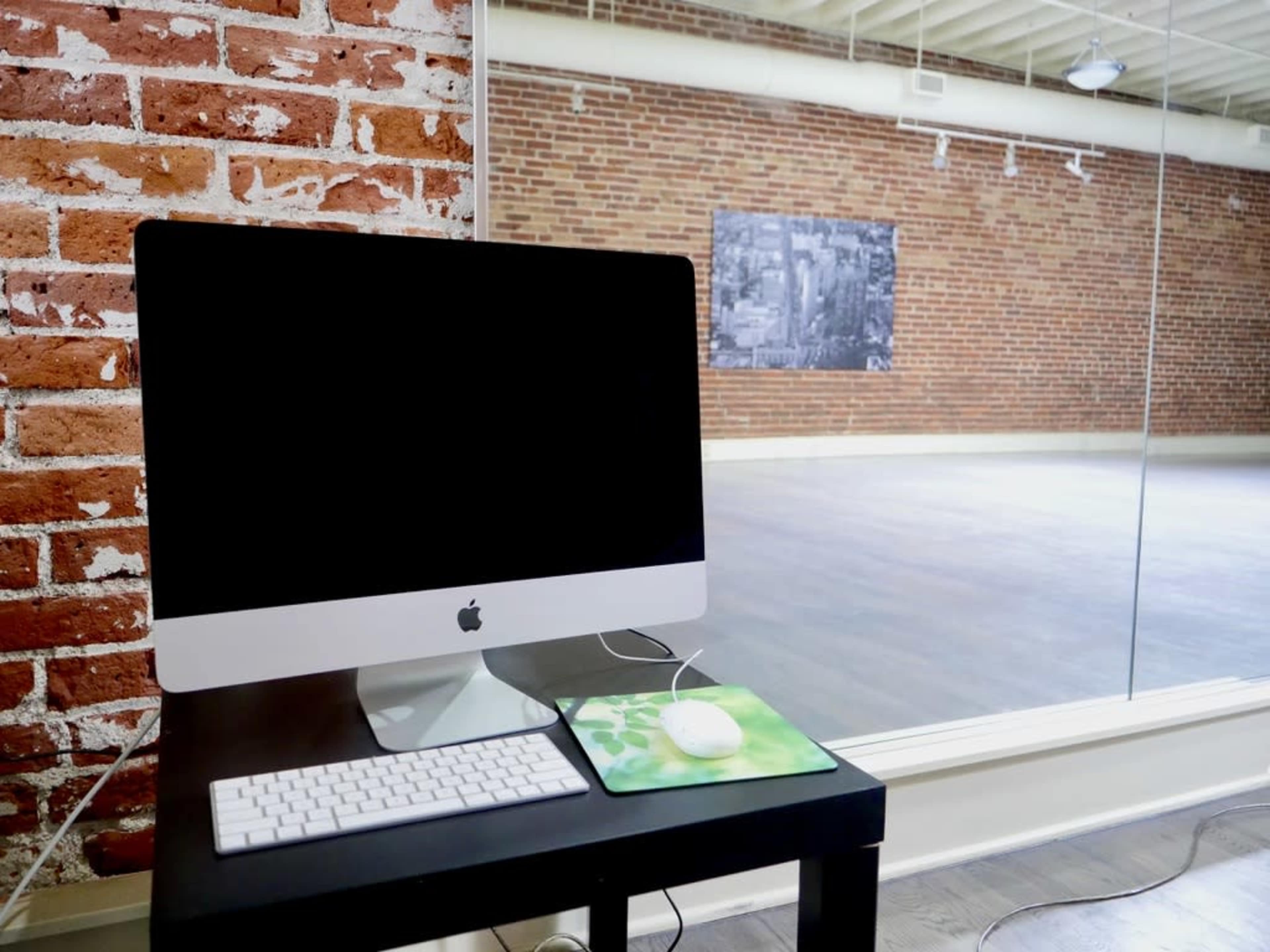 A computer and mouse are placed on a table in front of a large glass window, overlooking an empty studio space with exposed brick walls.