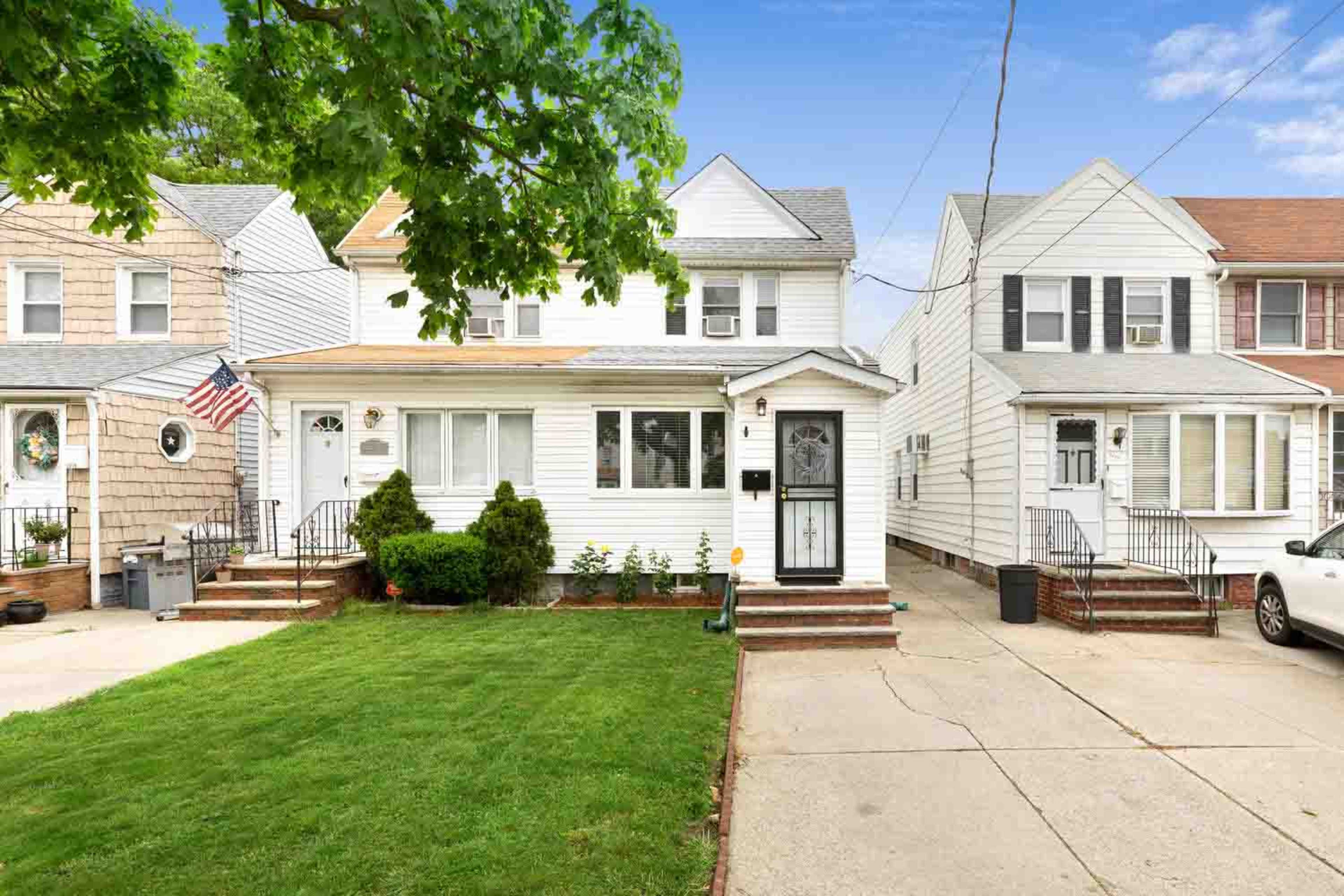A two-family house with a front lawn is flanked by two similar residential buildings, featuring a driveway and porch steps.