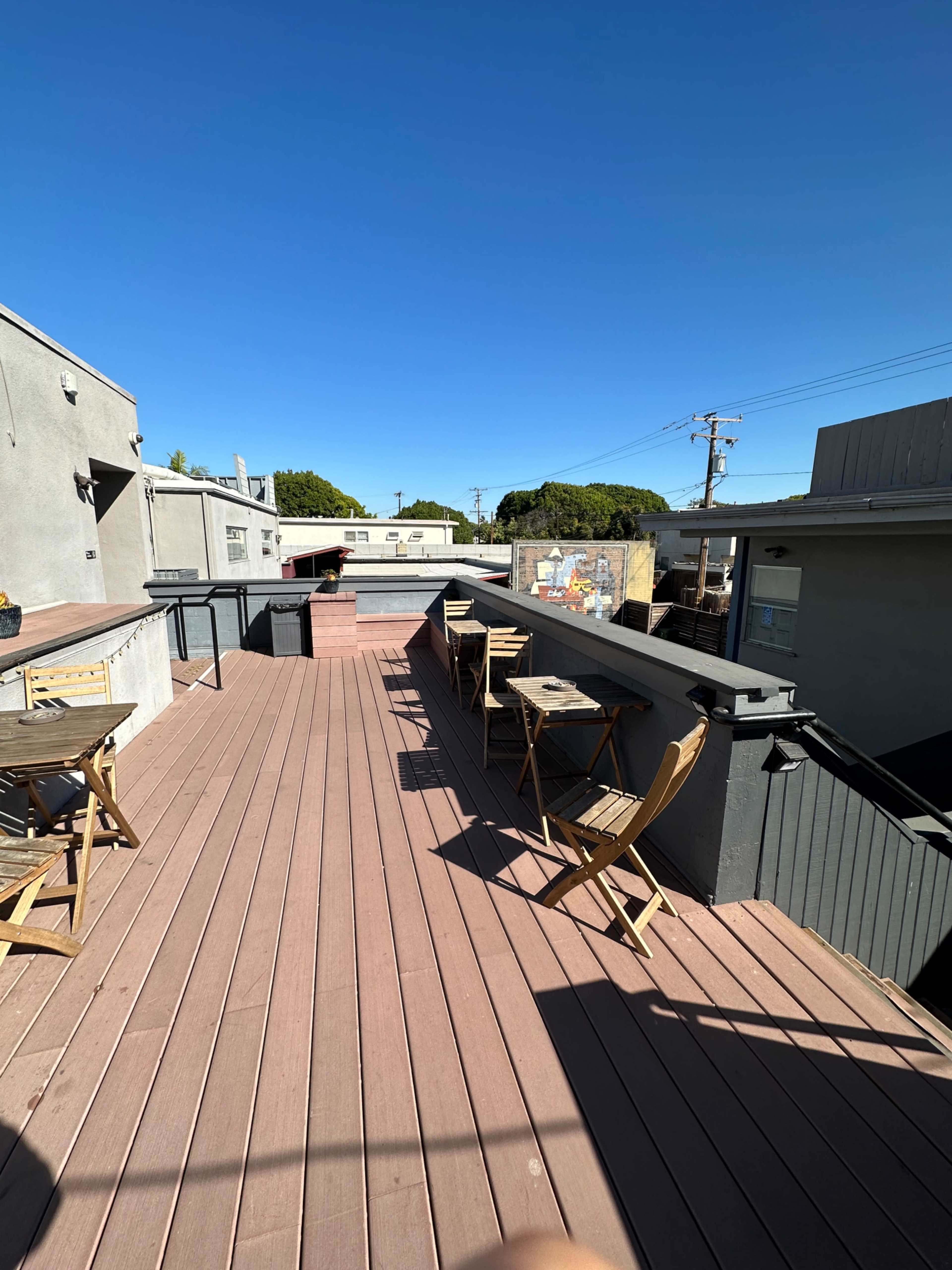 The image shows a rooftop deck with wooden flooring, several wooden chairs, and a railing, under a clear blue sky.