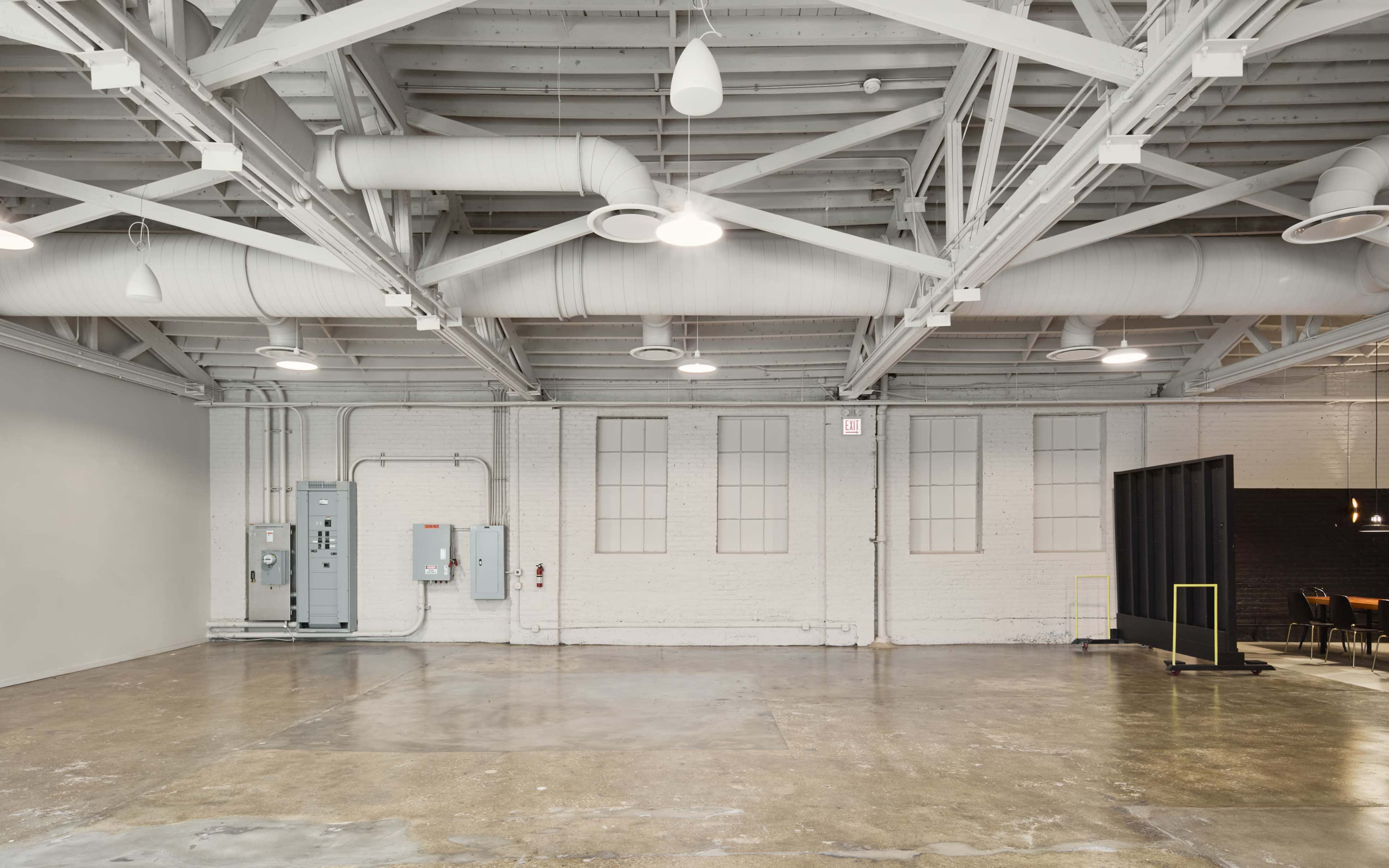 The image shows a spacious, empty industrial room with exposed beams, large windows, and polished concrete flooring.
