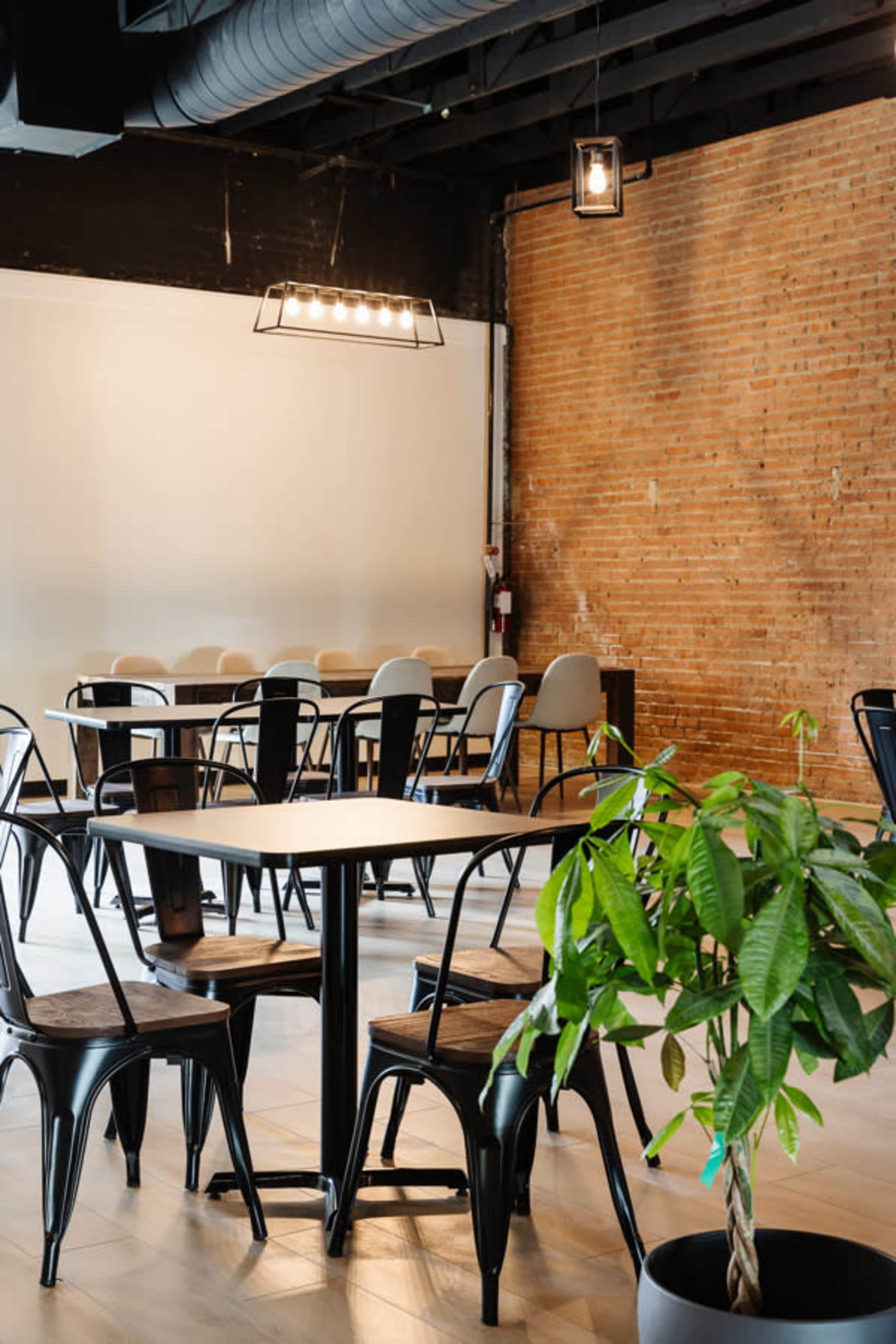 A modern dining area features a mix of metal and wooden chairs arranged around tables, with a brick wall and potted plant in the corner.