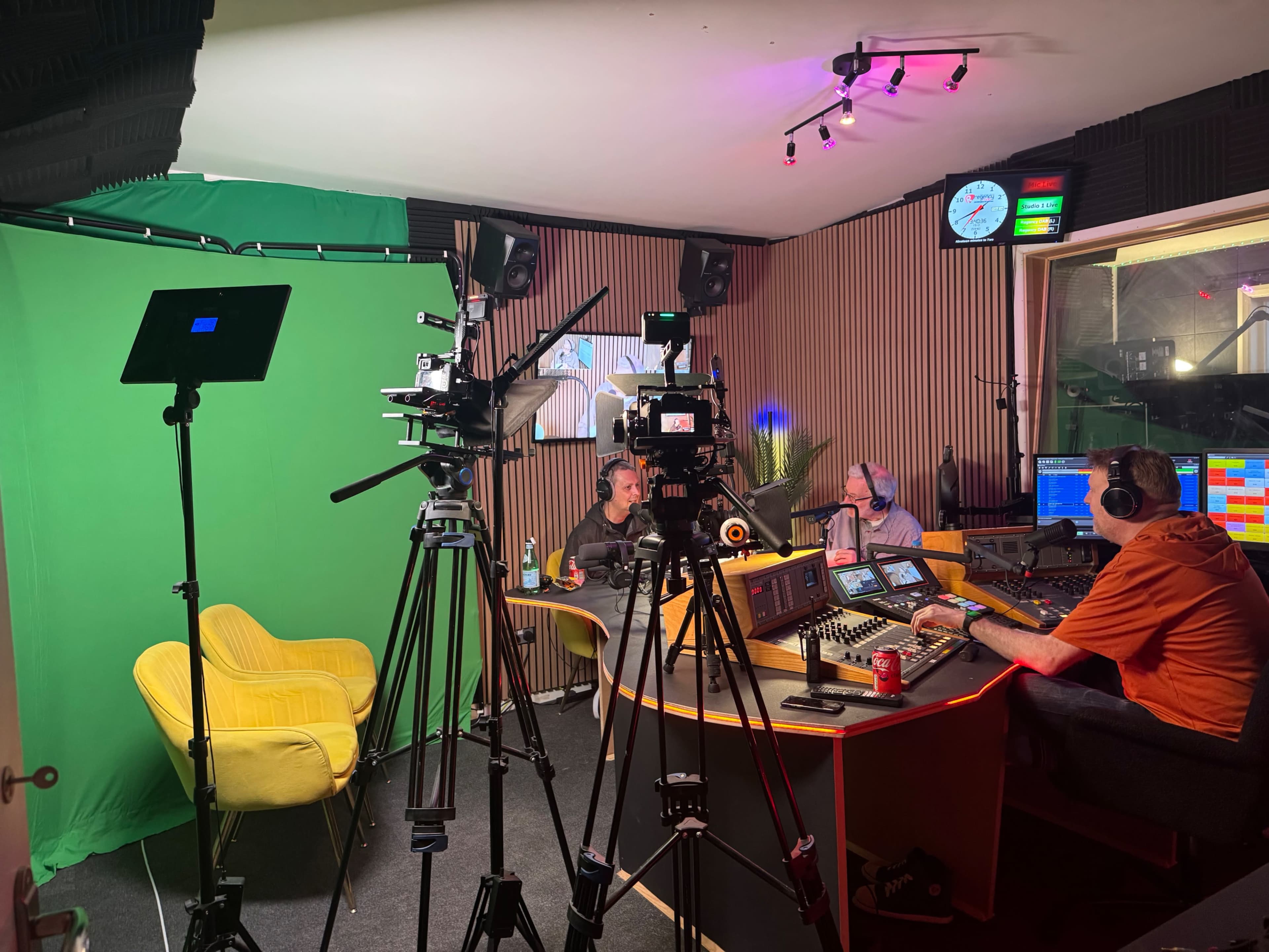 A recording studio is set up with cameras, a green screen, and three people engaged in a discussion at a central table.