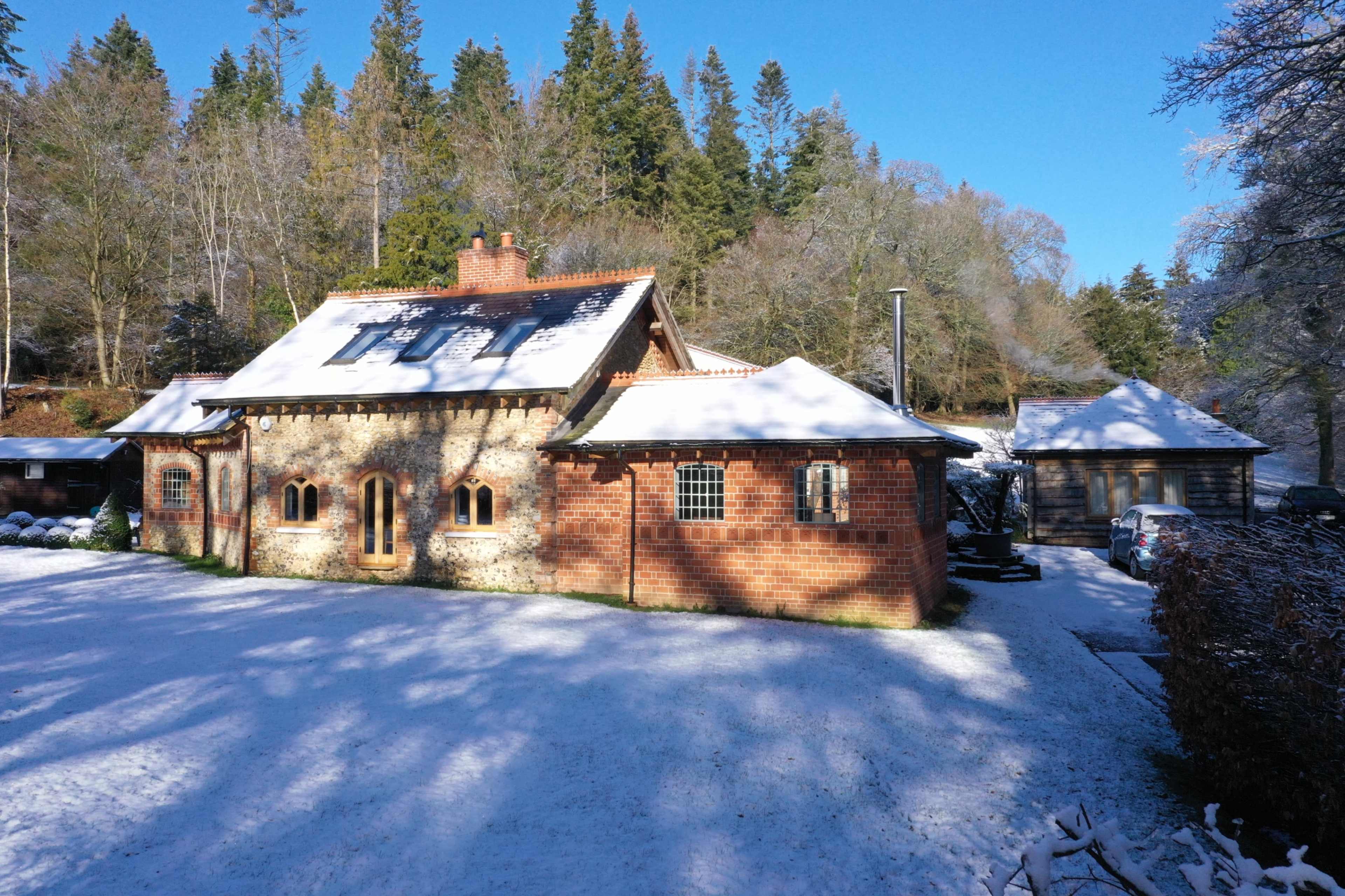 A brick and stone cottage is set against a snowy landscape, surrounded by trees and featuring a garage.