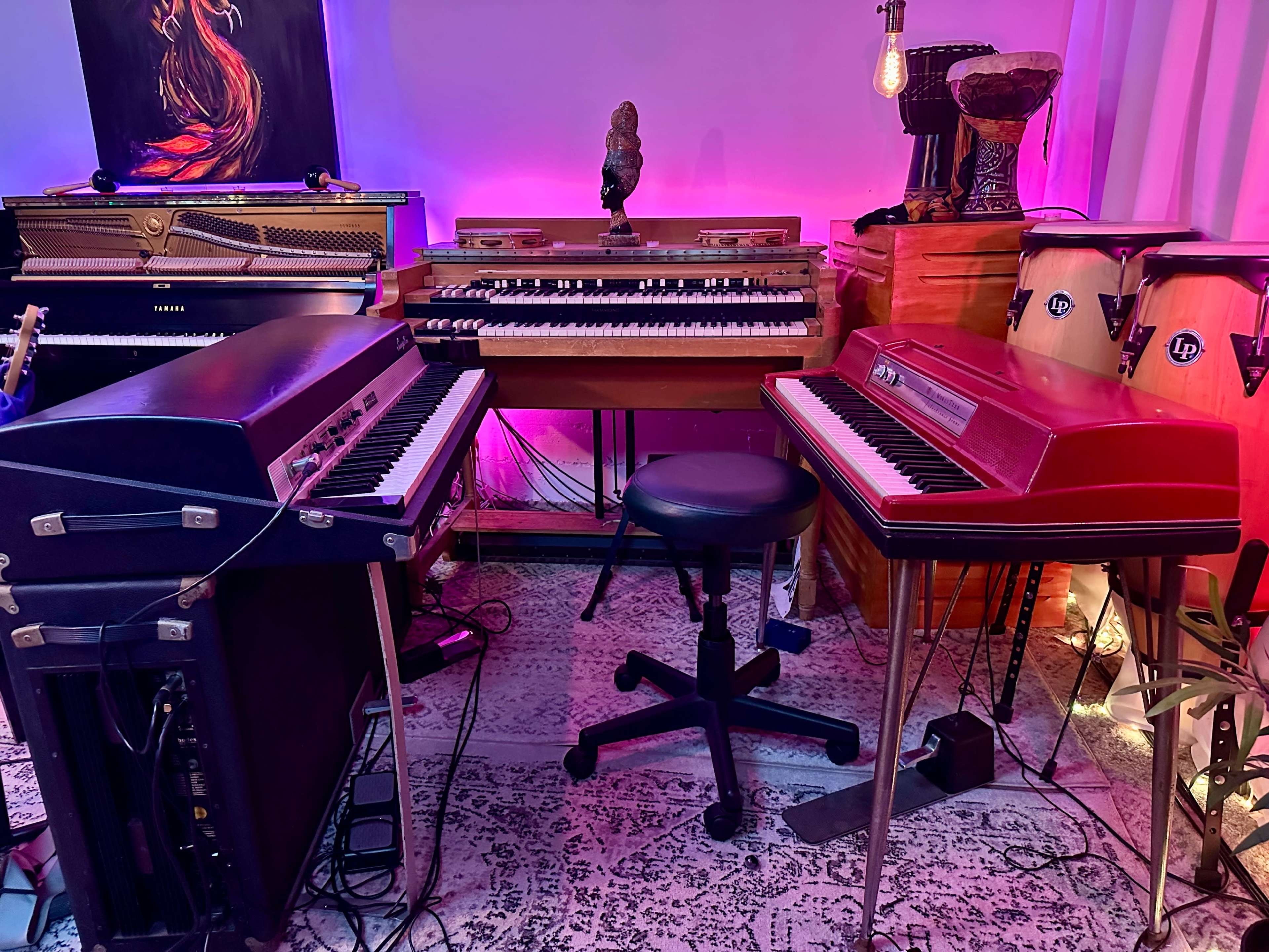 The image shows a music studio setup featuring two electric keyboards, a vintage organ, a drum, and various musical instruments, all illuminated by colorful lighting.