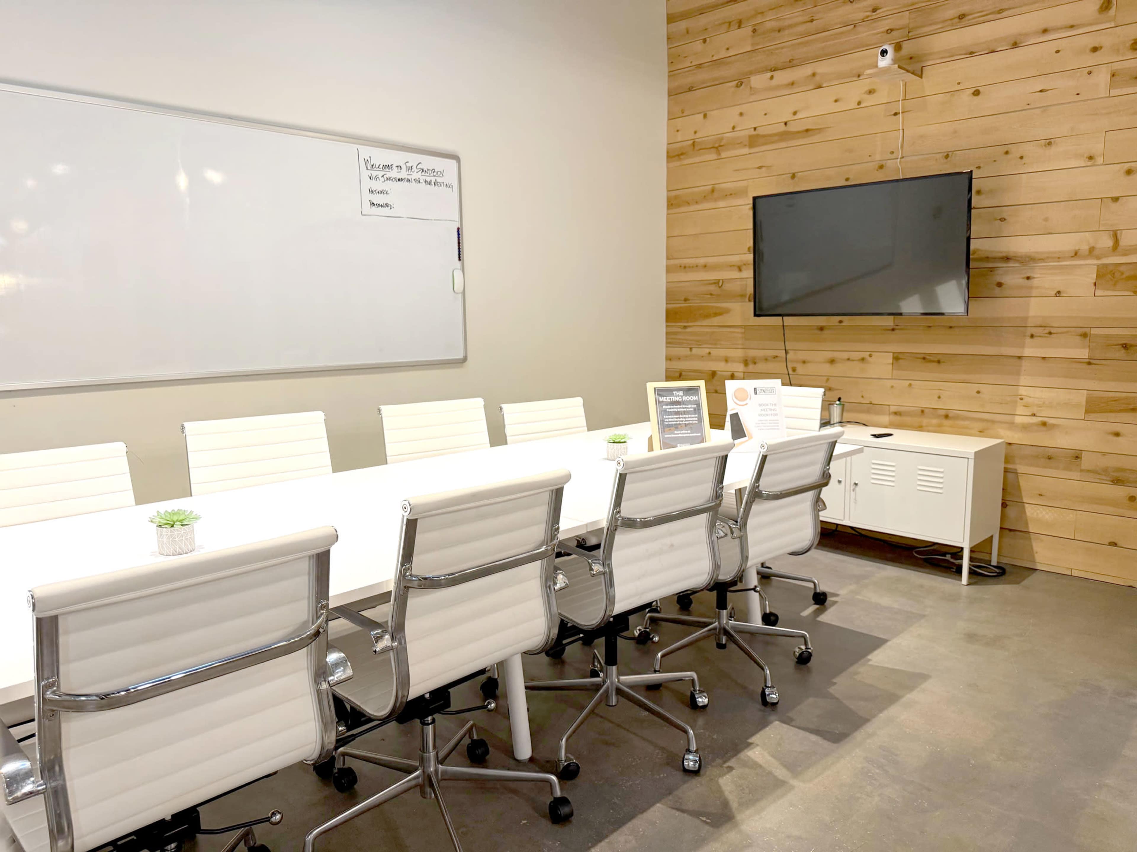 The image shows a modern conference room with a long white table, ergonomic chairs, a large television mounted on the wall, and wood-paneled accents.