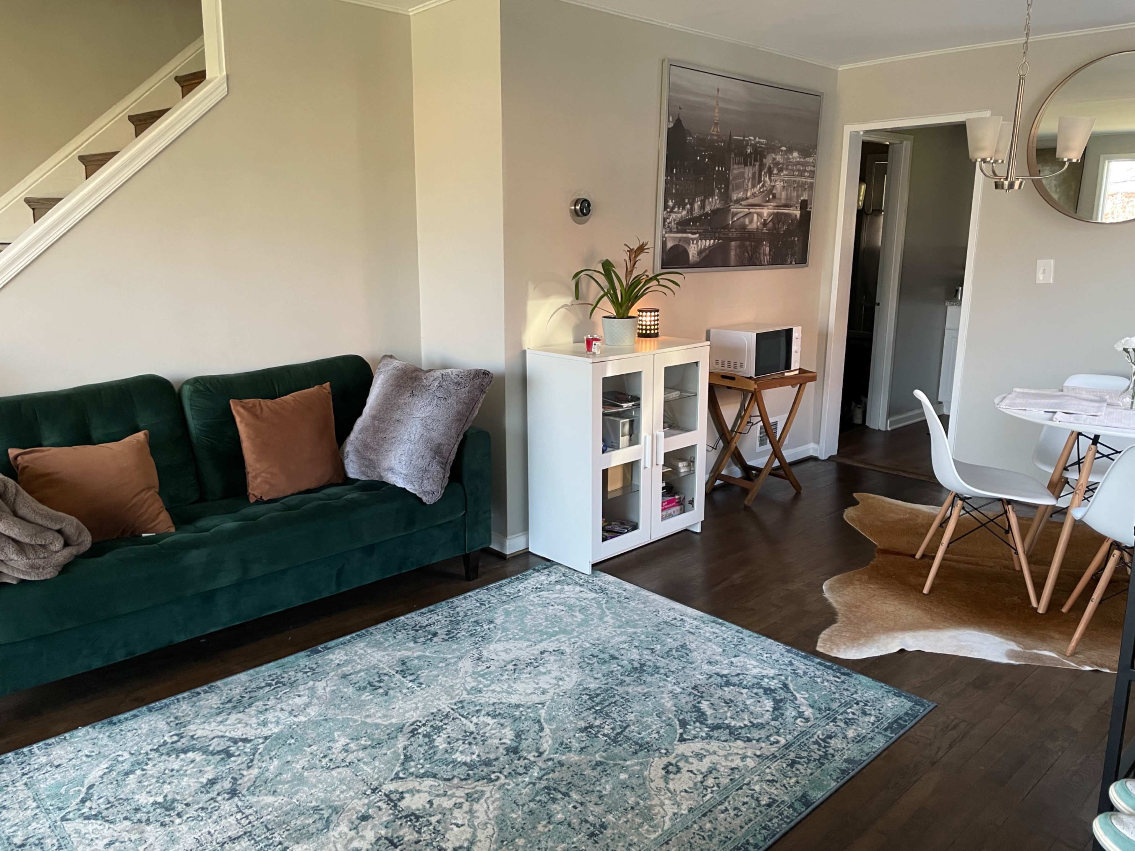 The room features a green sofa, a white shelving unit, a dining table with chairs, and a staircase in the background.