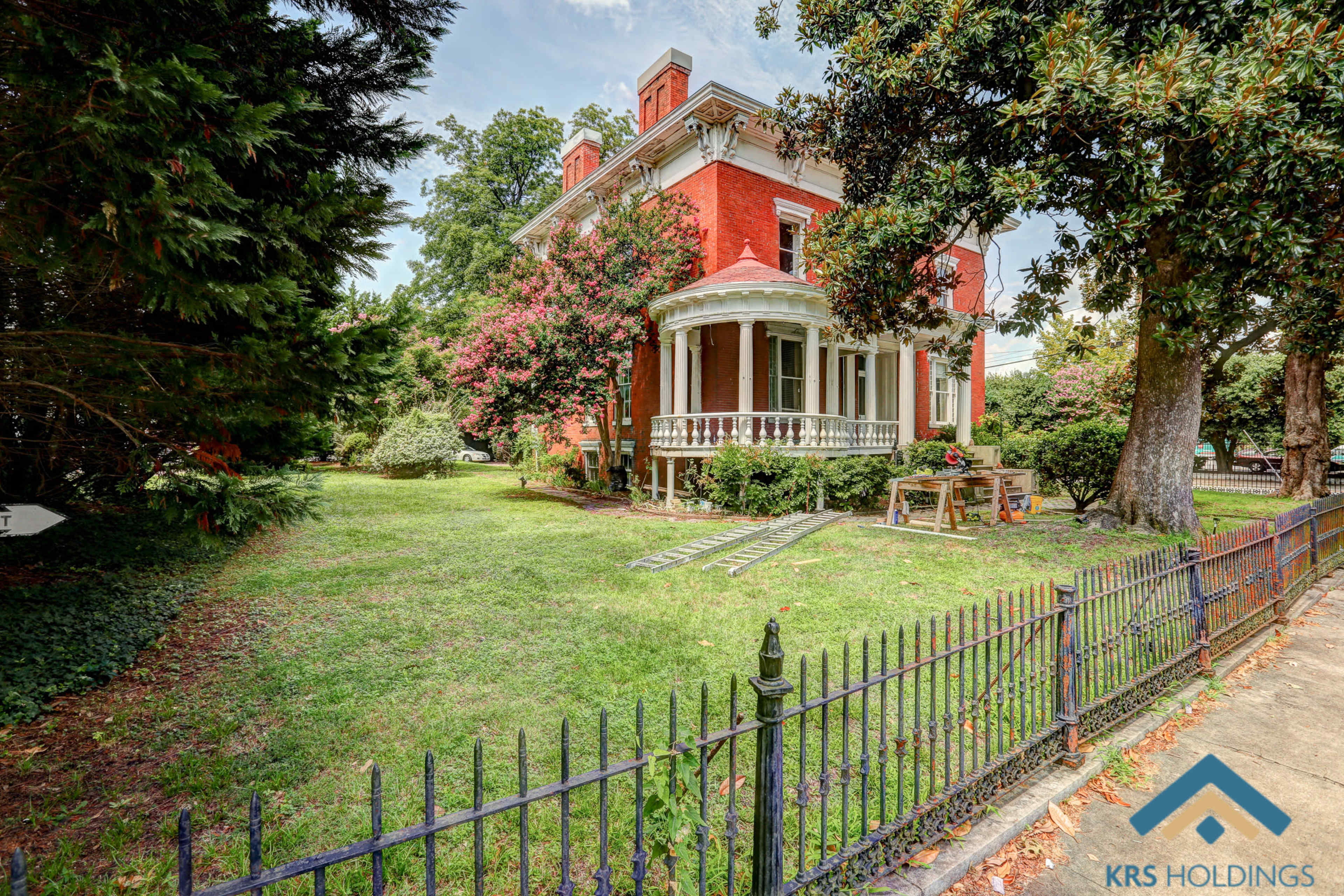 A red Victorian-style house with a front porch and well-maintained lawn is surrounded by trees and a black wrought-iron fence.