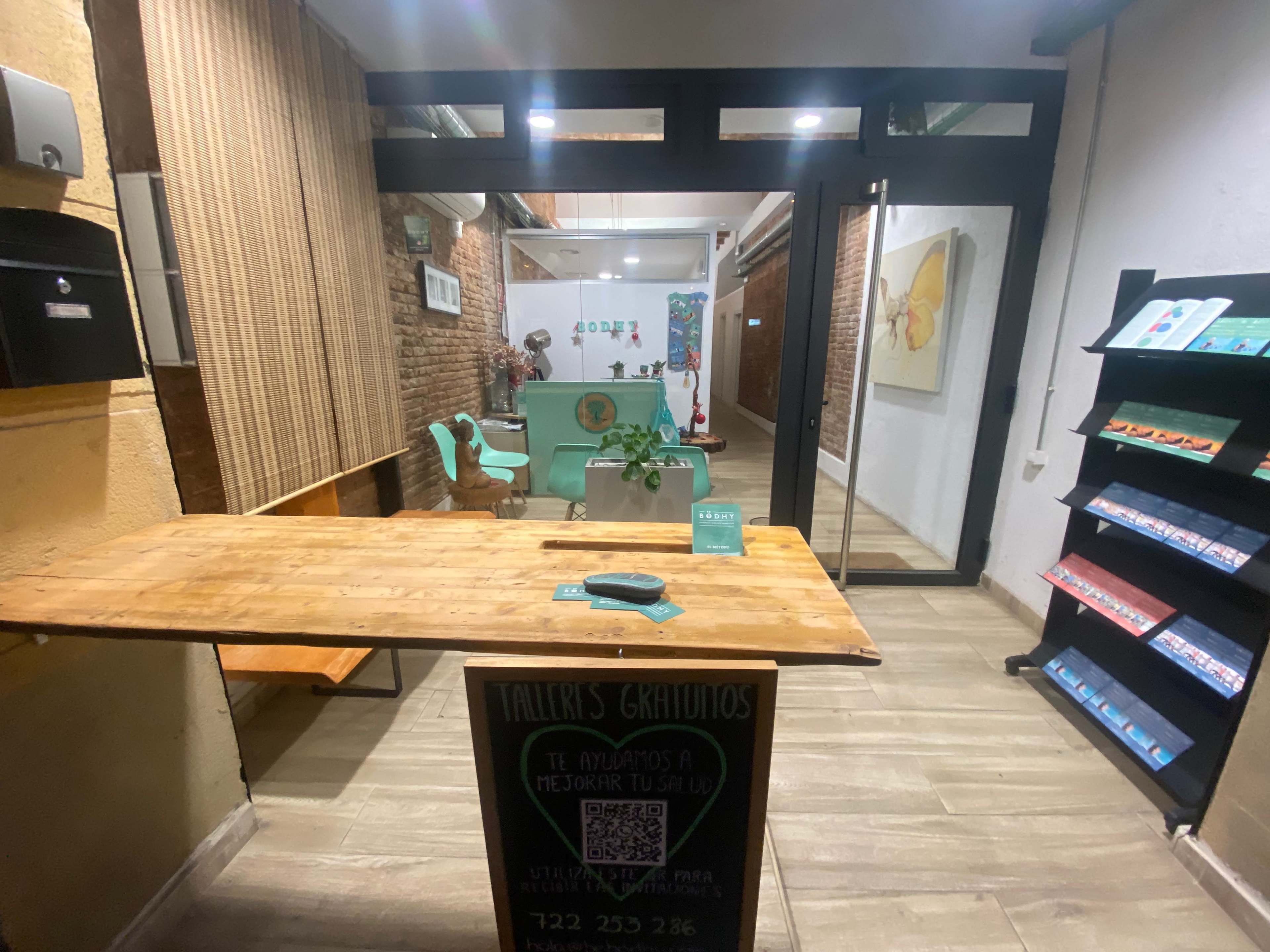 The image shows a modern reception area with wooden furniture, green seating, and a decorative wall featuring plants.
