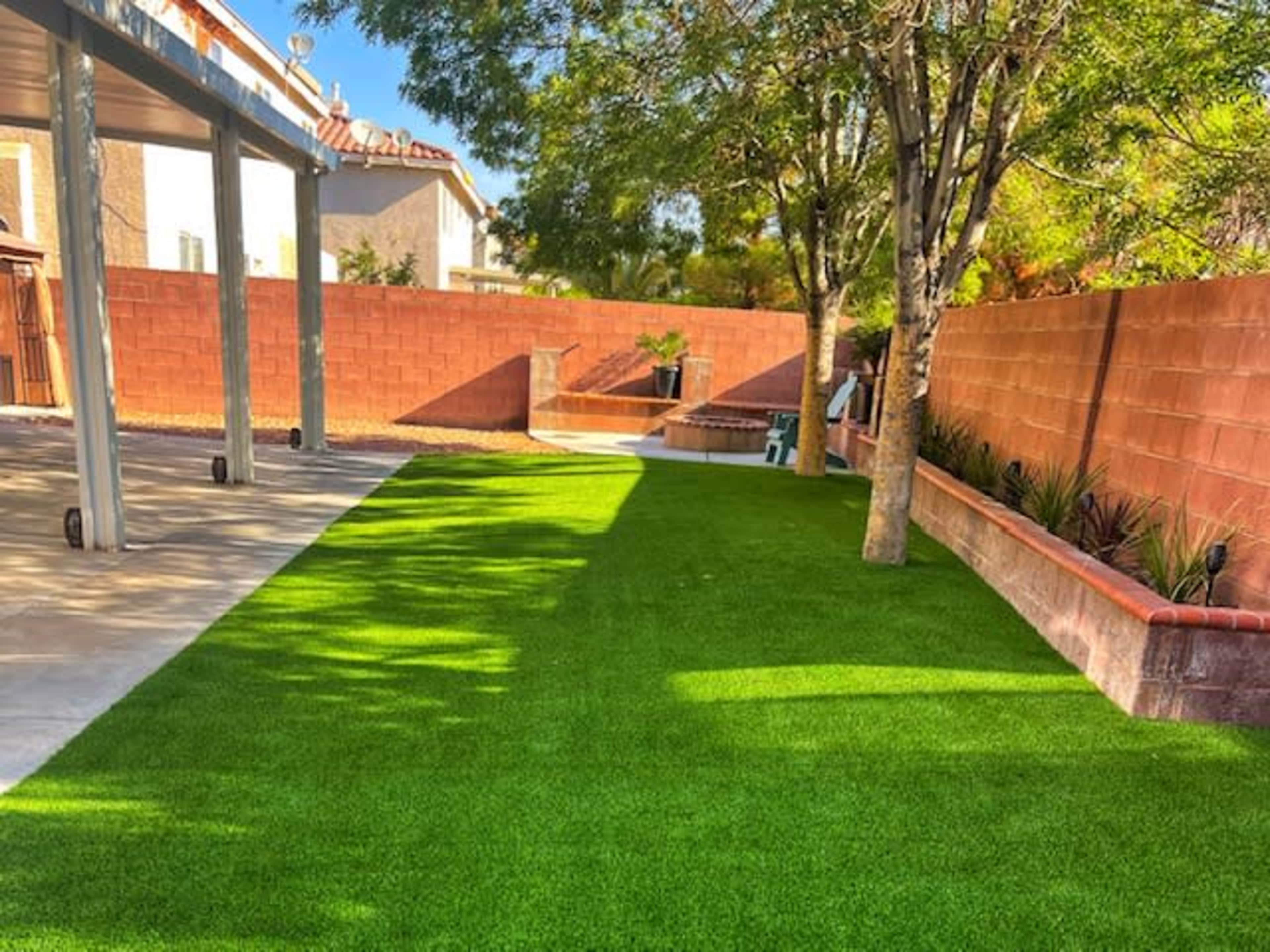 The image shows a well-maintained backyard with artificial turf, bordered by a red brick wall and shaded by trees.